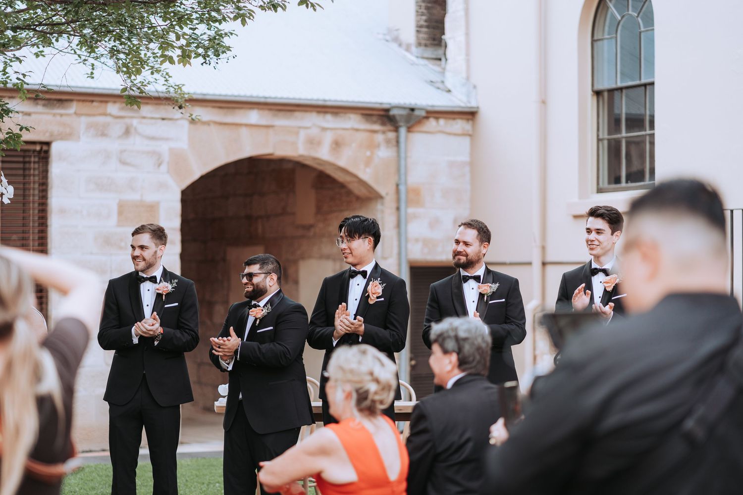 Wedding party and guests gathered outdoors near stone building during ceremony.