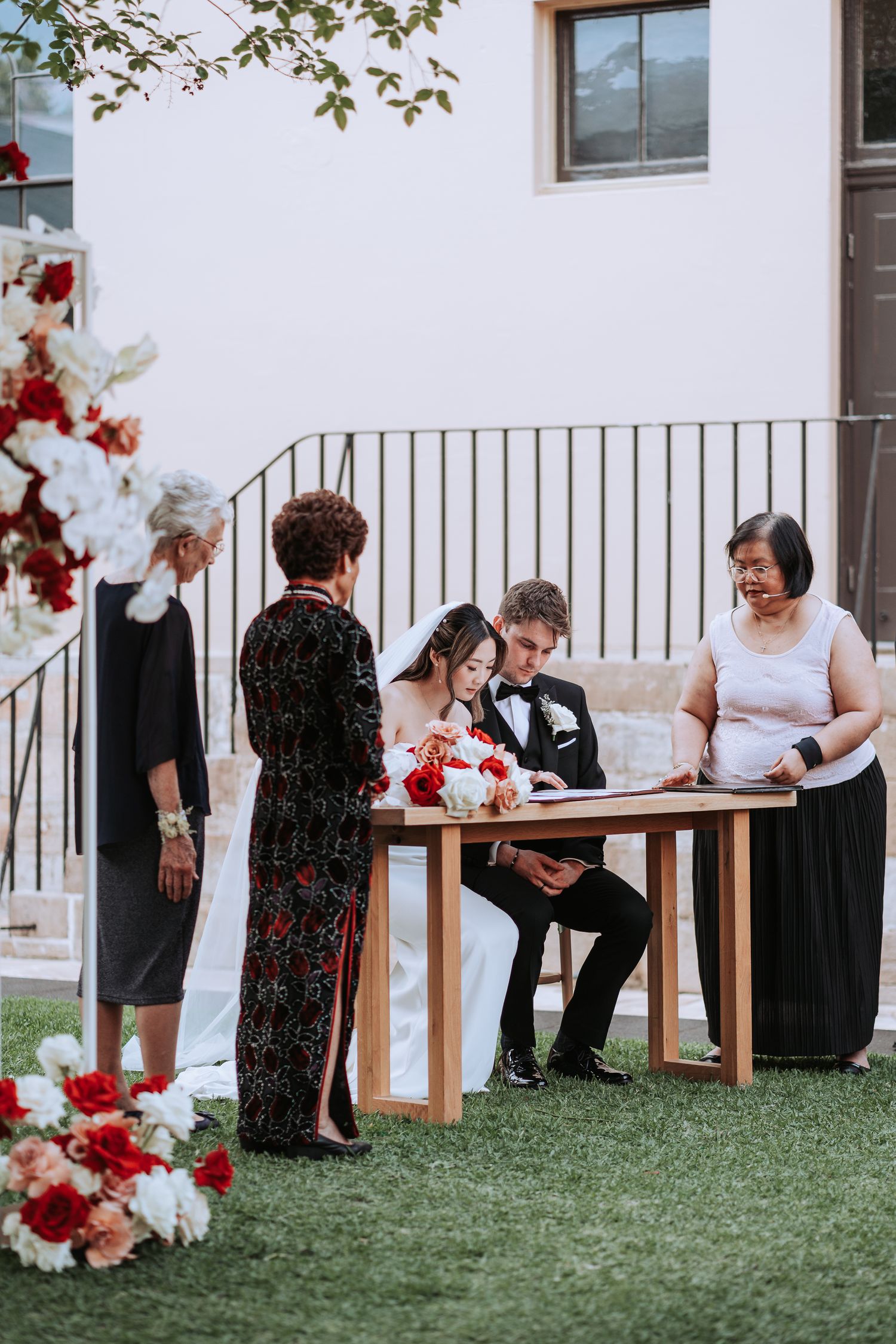 Marriage document signing ceremony with attendants gathered around wooden table outdoors.