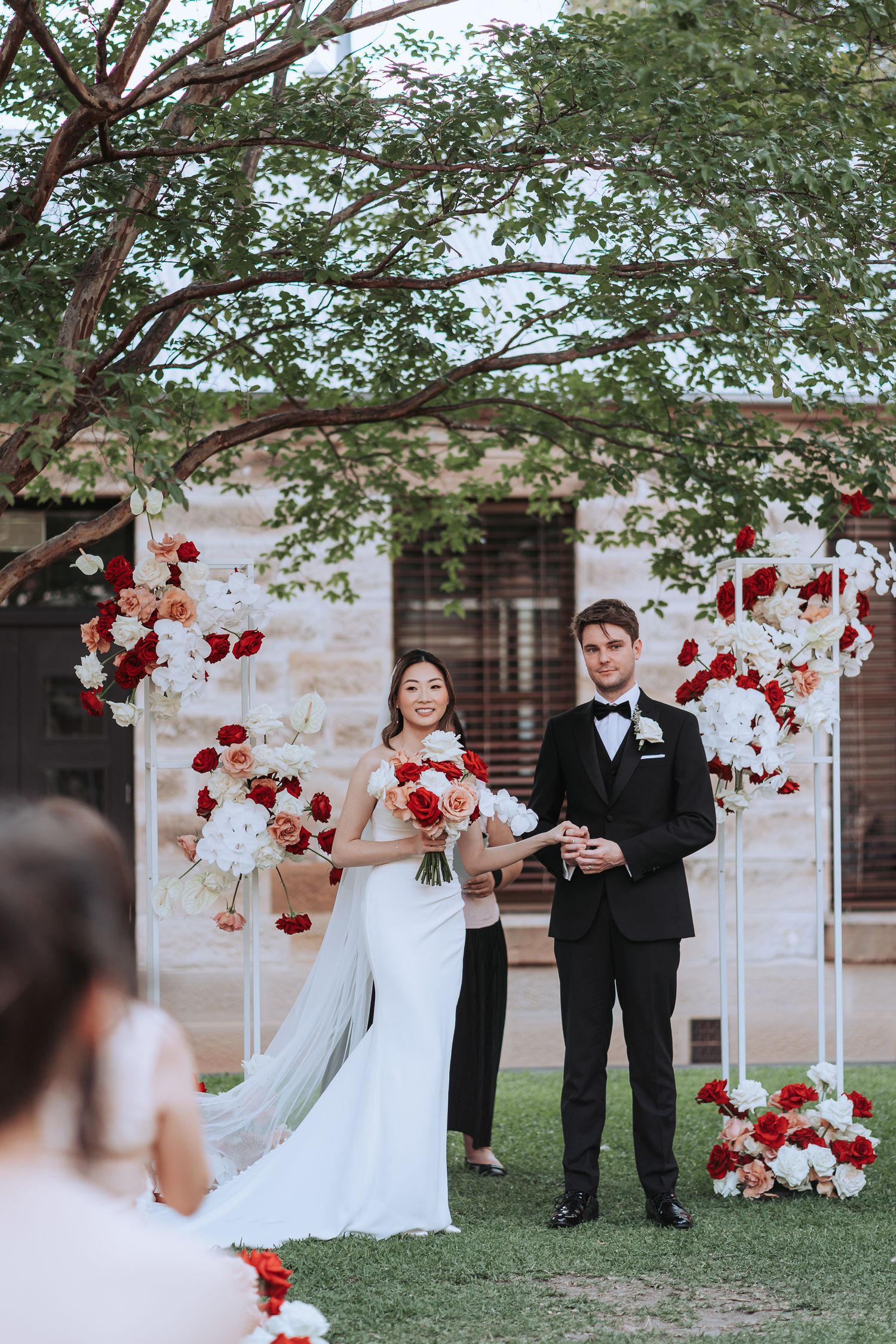 Newlyweds pose at outdoor ceremony with red and white floral arrangements and greenery overhead.