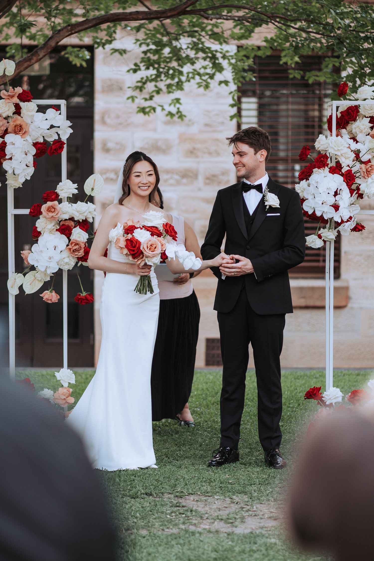 A wedding ceremony with a couple standing under floral red and white arch decor against a stone wall.