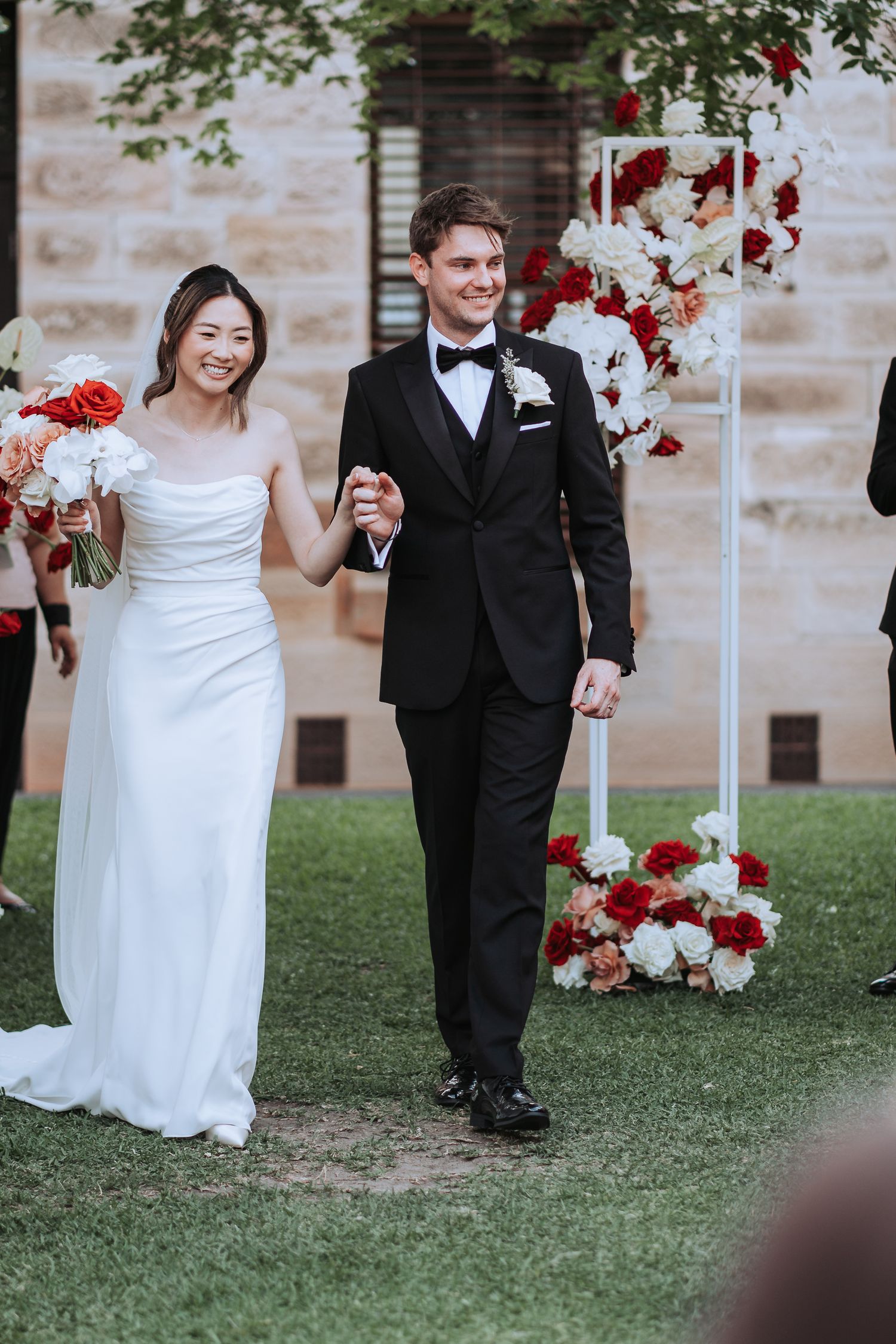 Happy newlyweds walking hand in hand past elegant red and white floral arrangements.