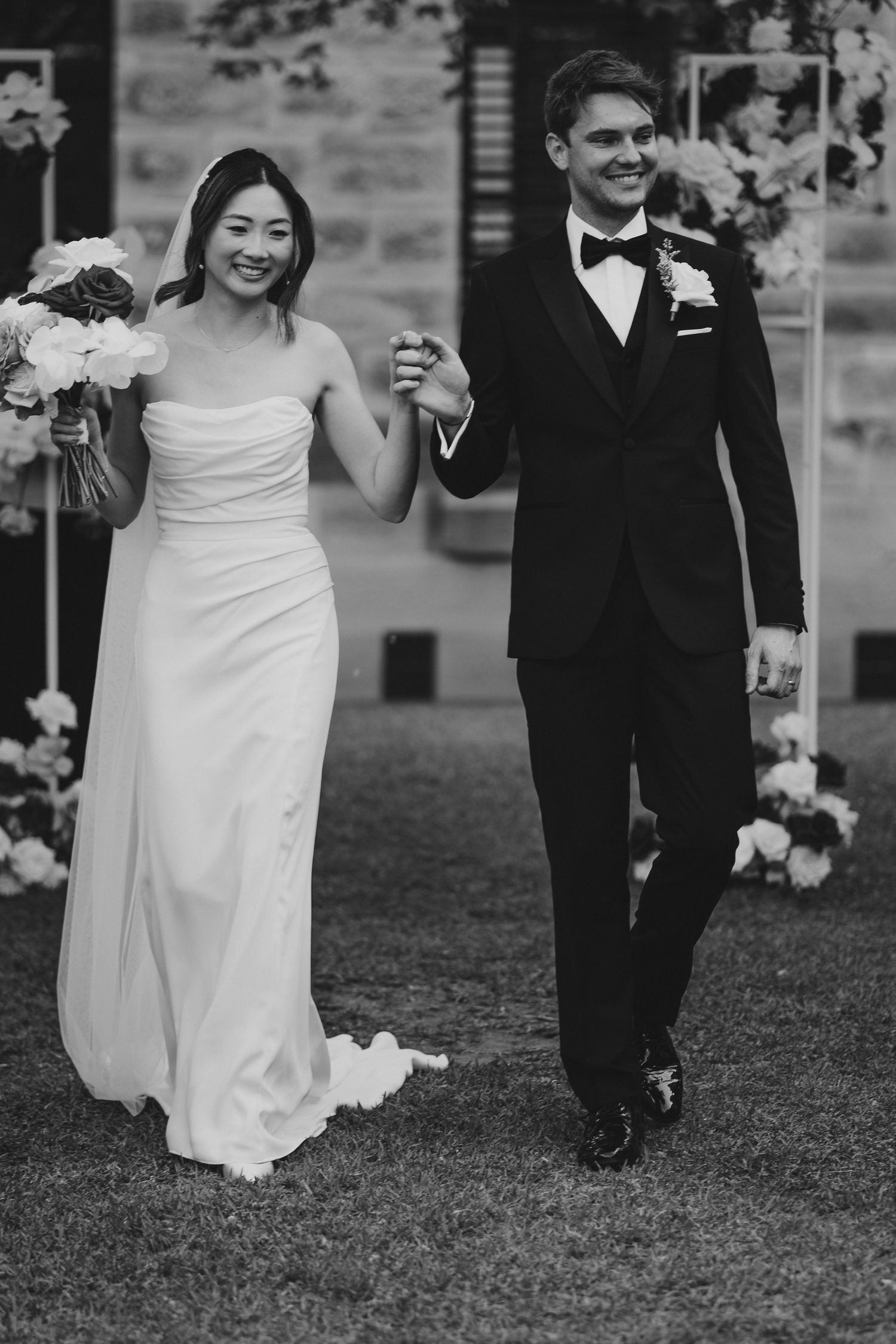 Black and white photo of newlyweds holding hands while walking on grass after ceremony.