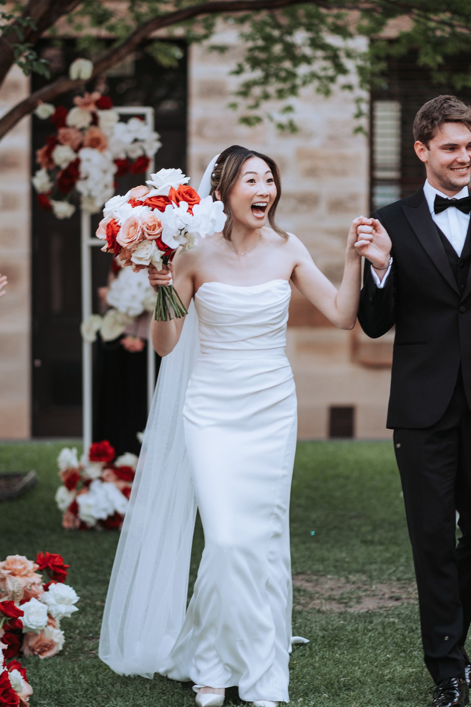 Joyful wedding couple walks together with red and pink bouquet surrounded by floral decorations.