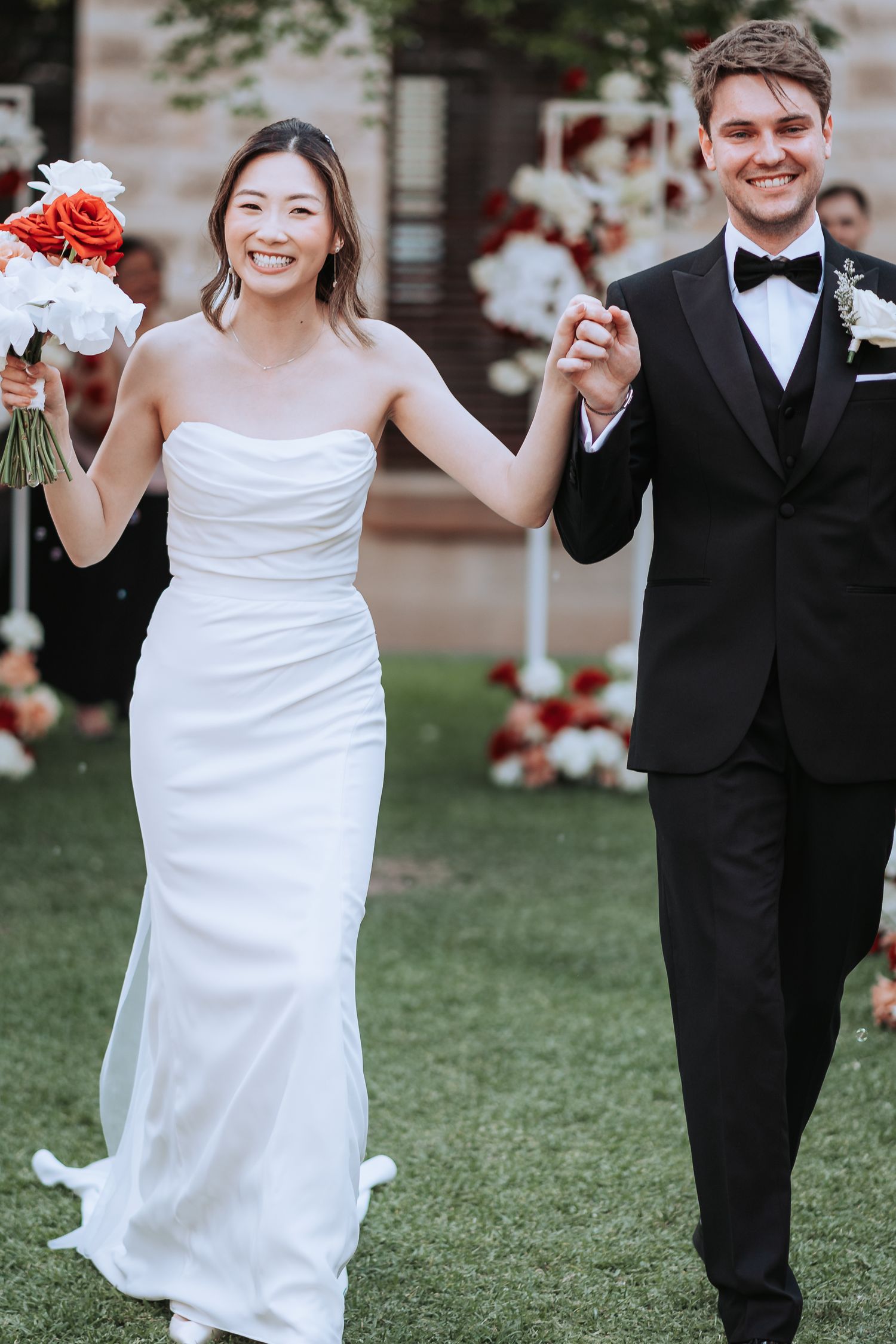 Newlyweds share joyful moment walking together with red and white bouquet on wedding day.