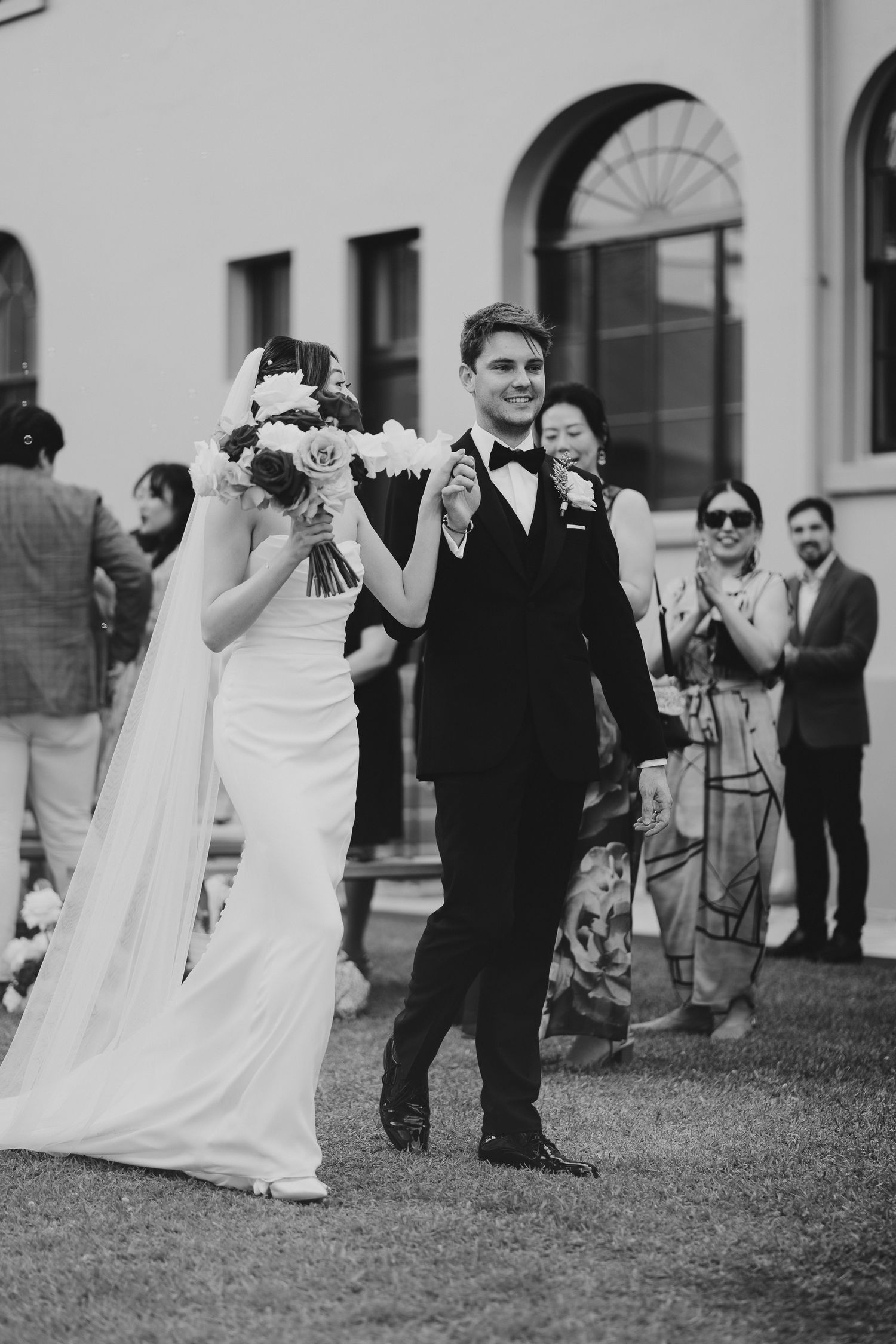 Black and white photo shows wedding couple walking past guests during outdoor celebration.