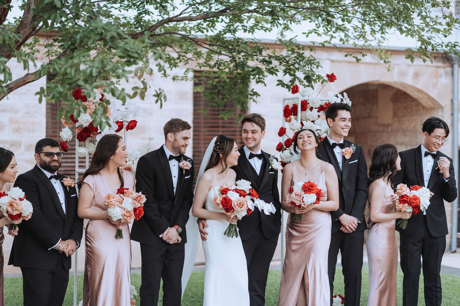 Wedding party posed outdoors with pink bridesmaid dresses and red floral arrangements.