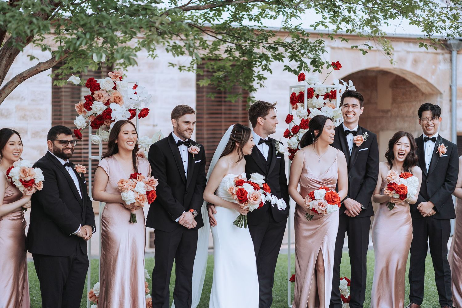 Wedding party lineup with bridesmaids in pink dresses and groomsmen in black tuxedos.
