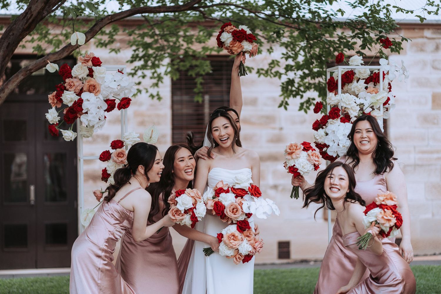 Bridesmaids in pink dresses laughing with bride holding red and white bouquets.