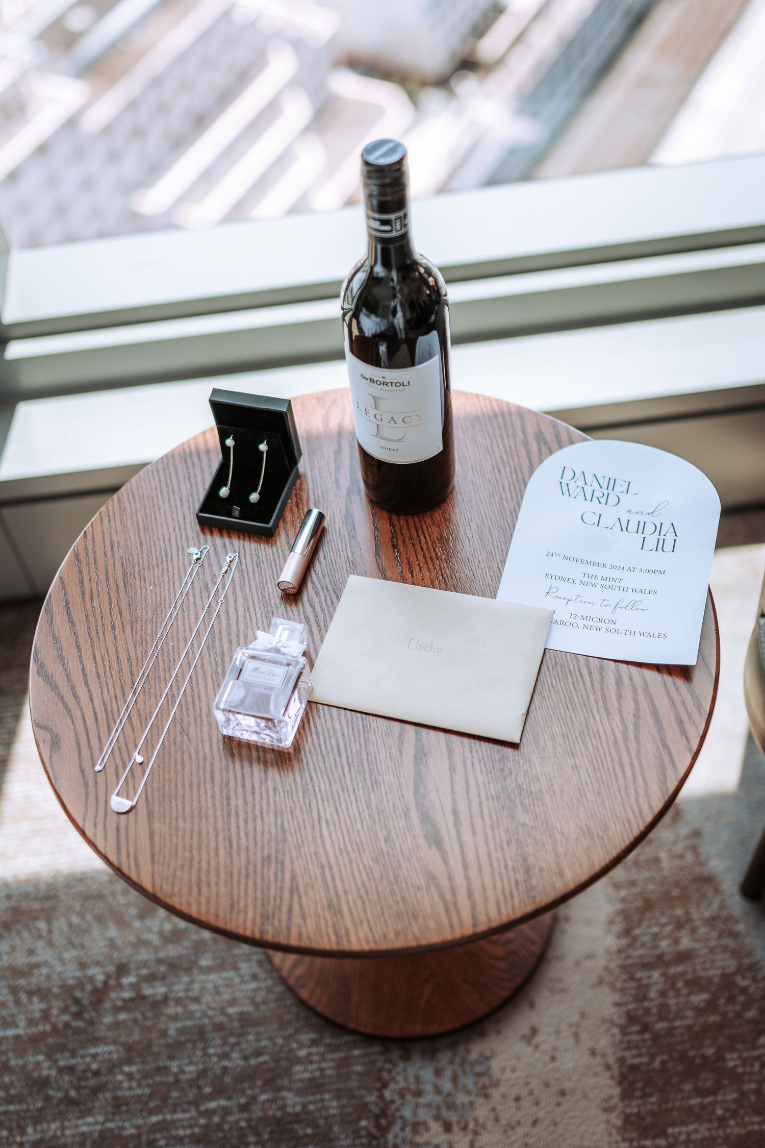 Wine bottle, jewelry box and stationery arranged on wooden side table by window.