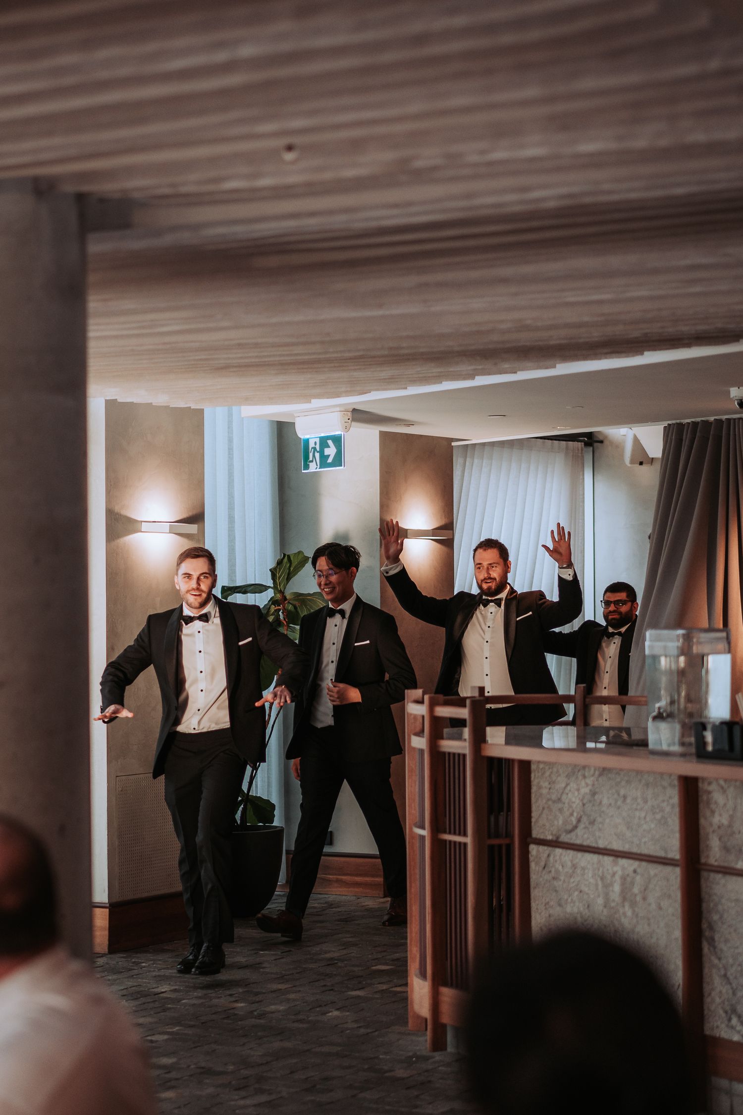 Groomsmen make a grand entrance at wedding reception wearing black tuxedos in modern venue space.