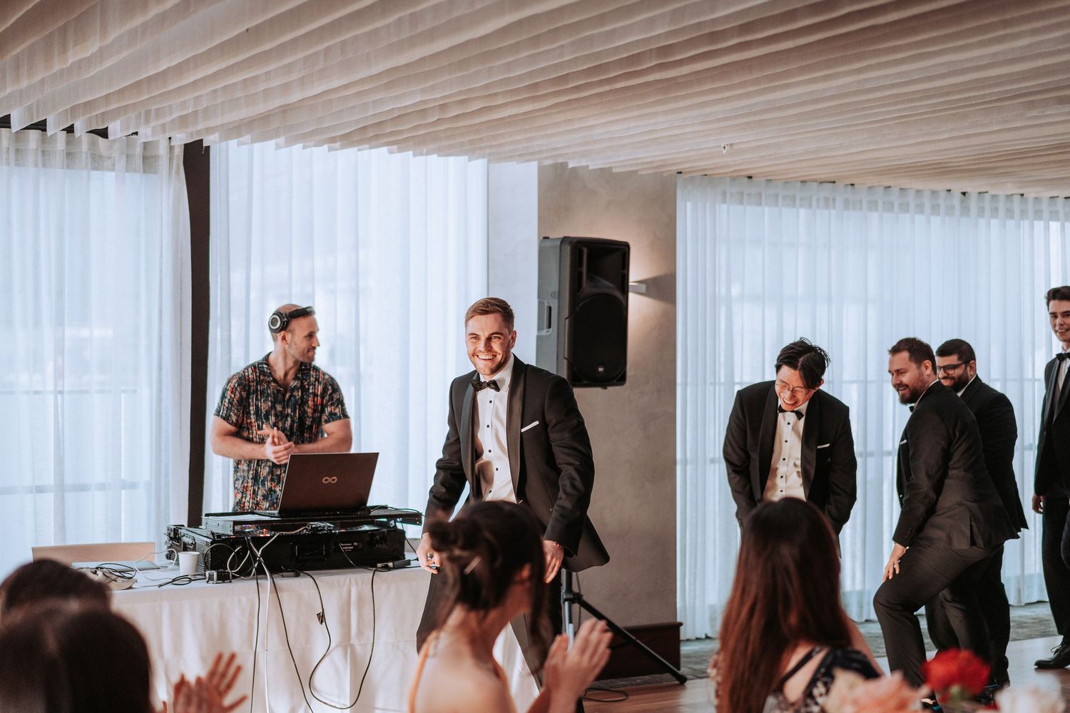 Wedding party dances while DJ performs at modern reception venue with white curtains.