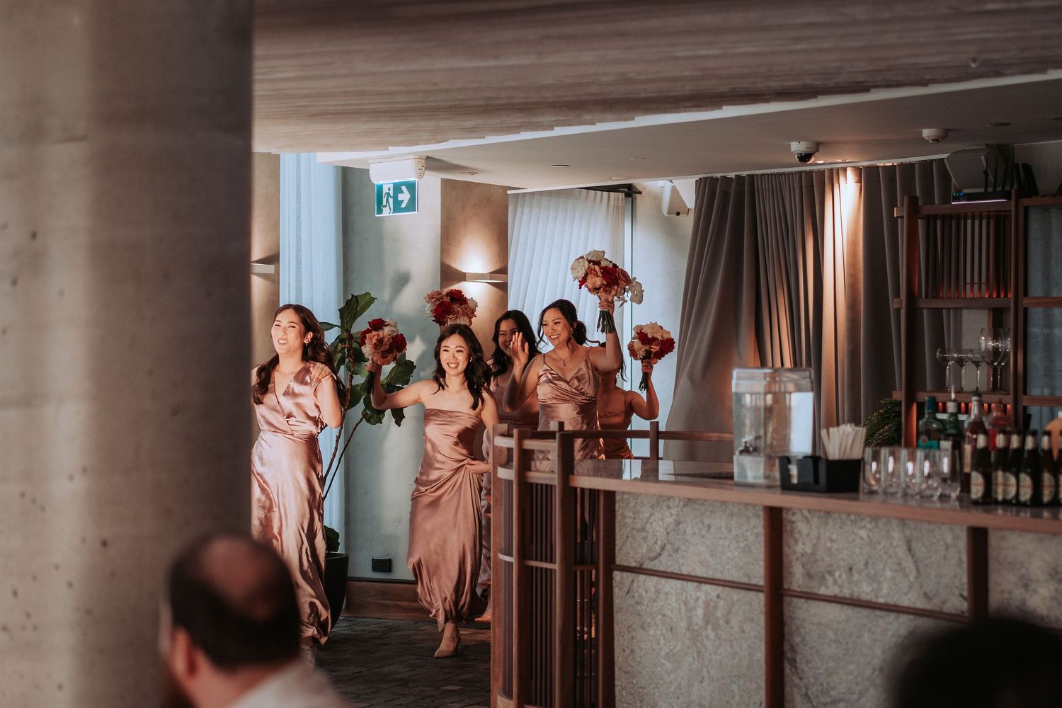 Wedding party members in pink dresses gather near bar area during reception celebration.