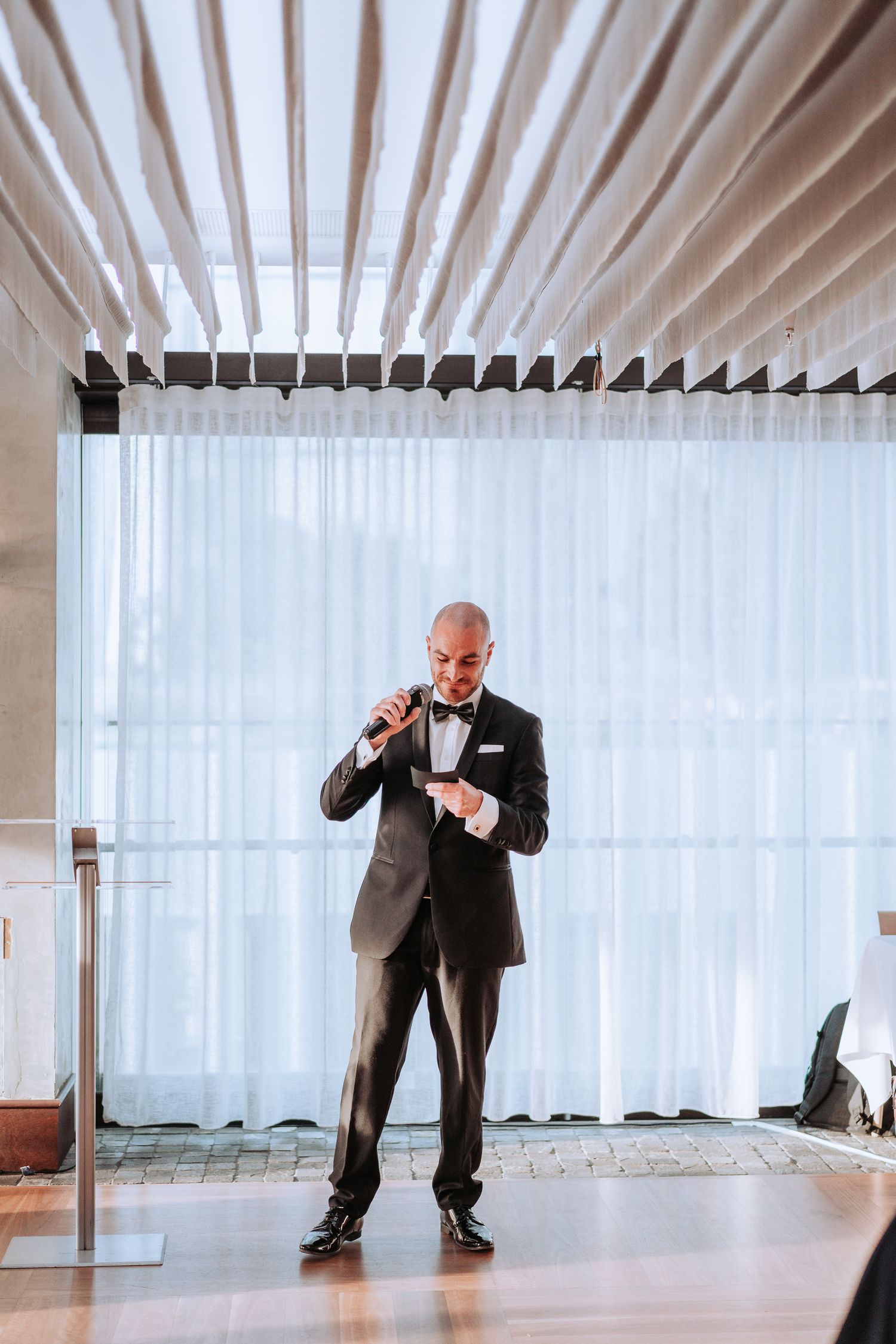 Person in formal suit delivers speech at modern wedding venue with white backdrop.