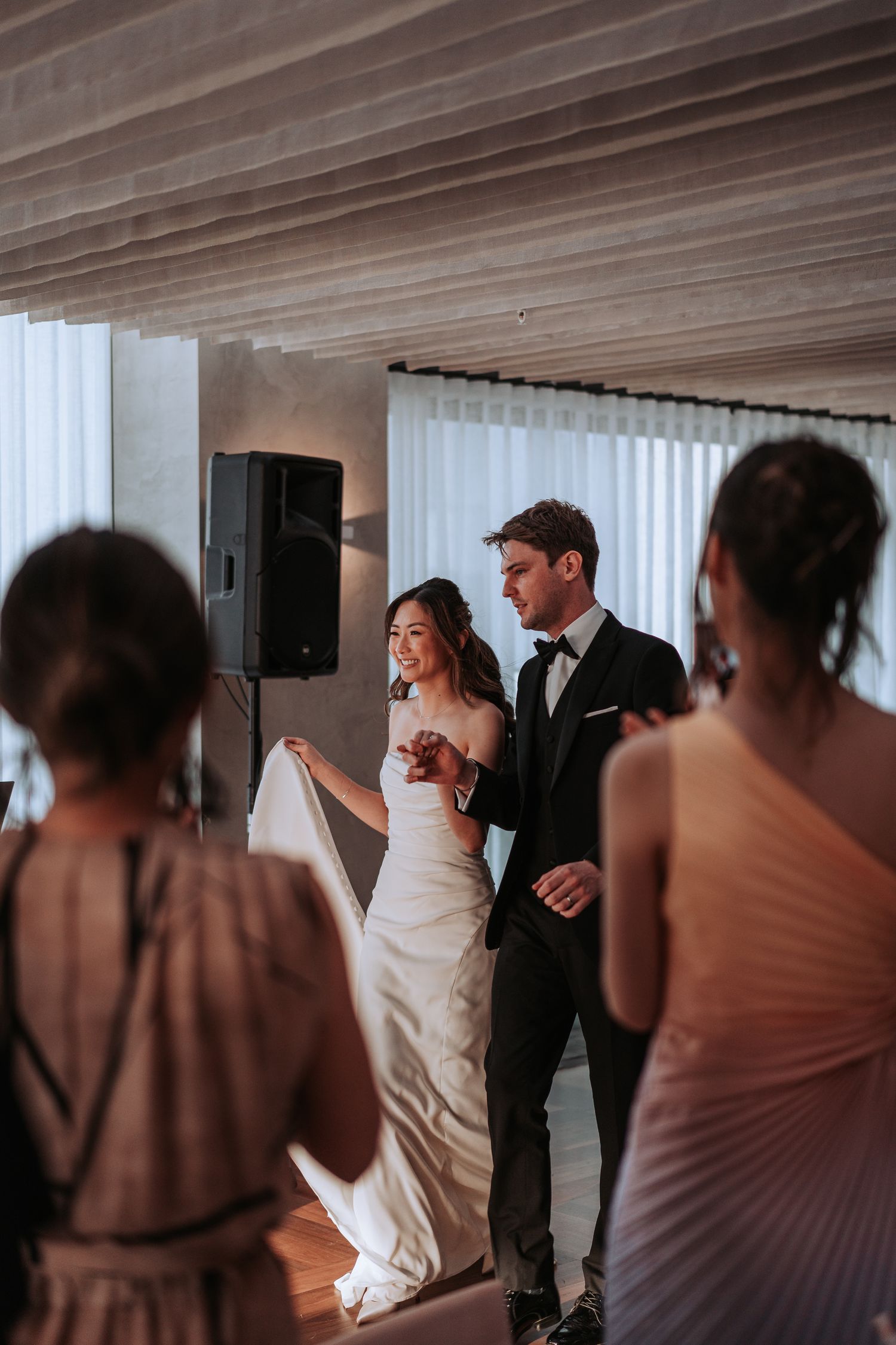 Newlyweds dance together during their wedding reception in a modern venue with white drapes and wooden ceiling details.