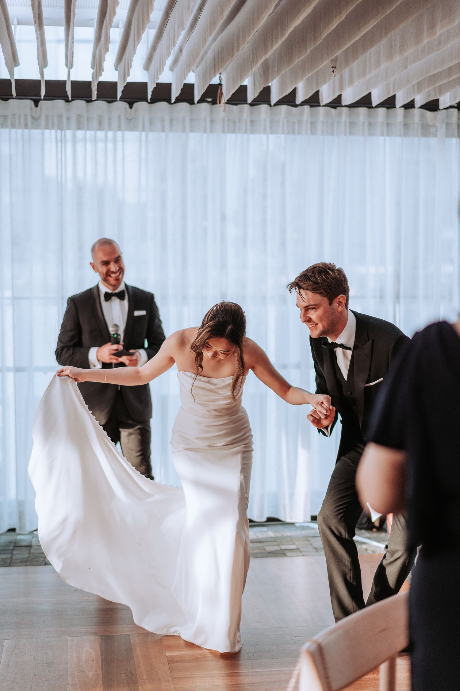 Wedding couple dancing while guest looks on at modern reception venue with white curtain backdrop.