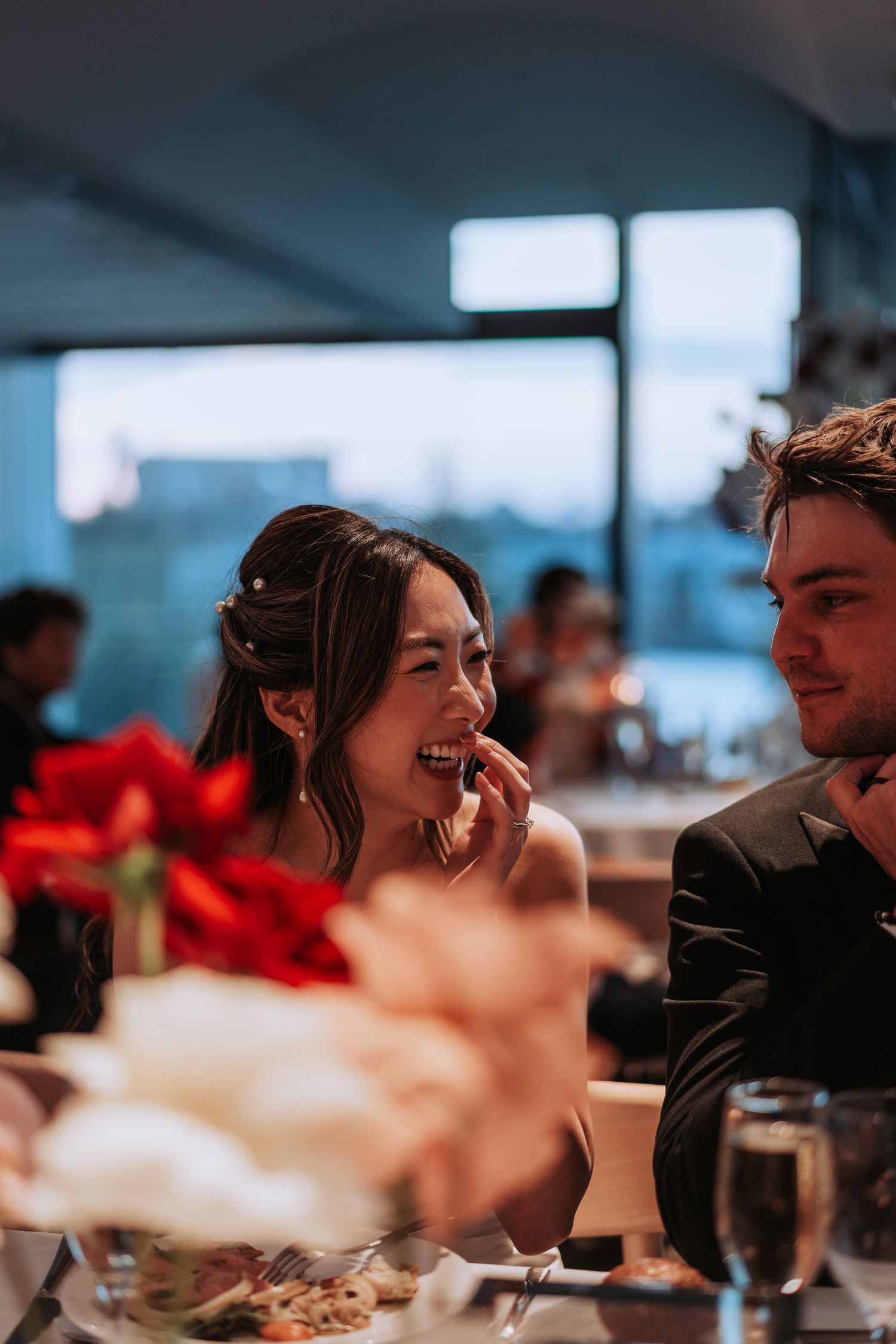 Candid moment of laughter during wedding reception dinner with red floral decorations.