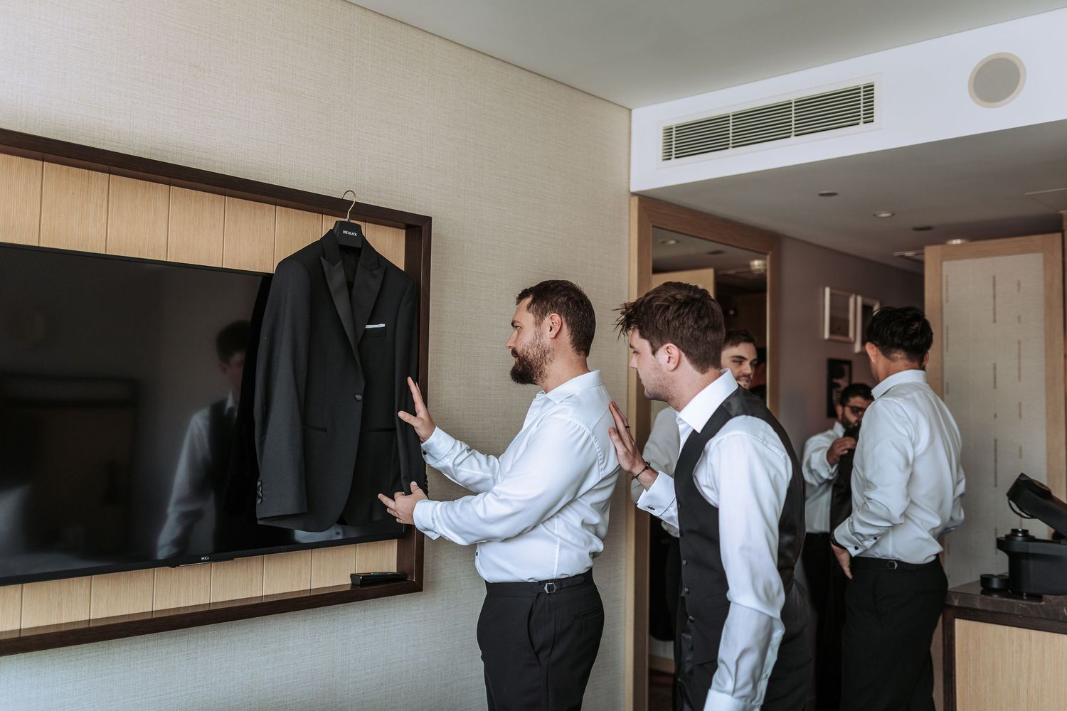 Groomsmen getting dressed in a hotel room with formal attire hanging on wall hooks.