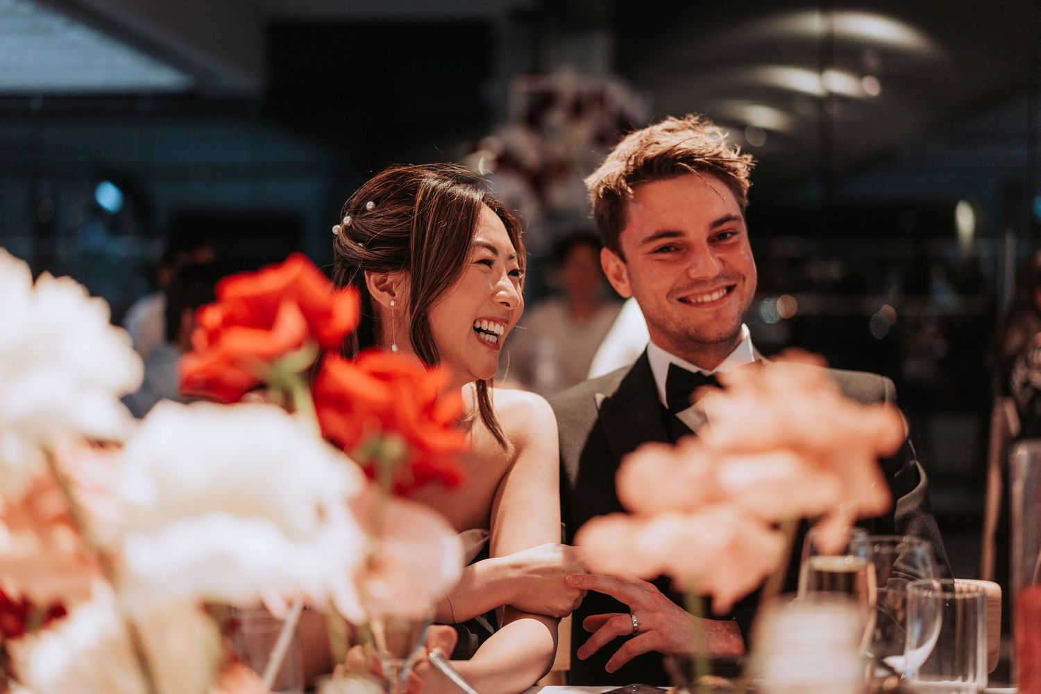 Newlyweds share a candid moment of laughter at their reception surrounded by coral and white flowers.