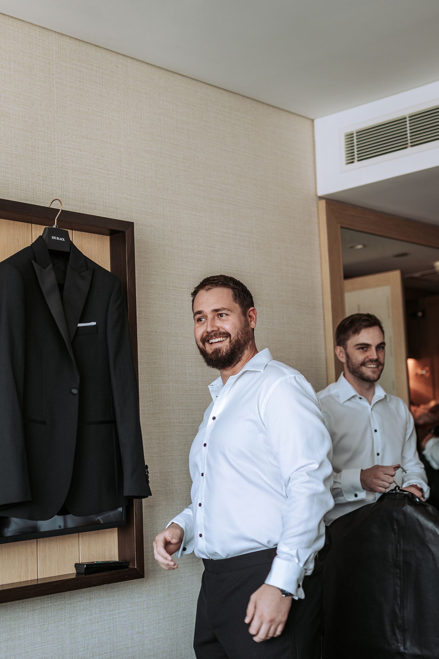 Wedding party members smiling while getting ready in front of hanging formal suits.