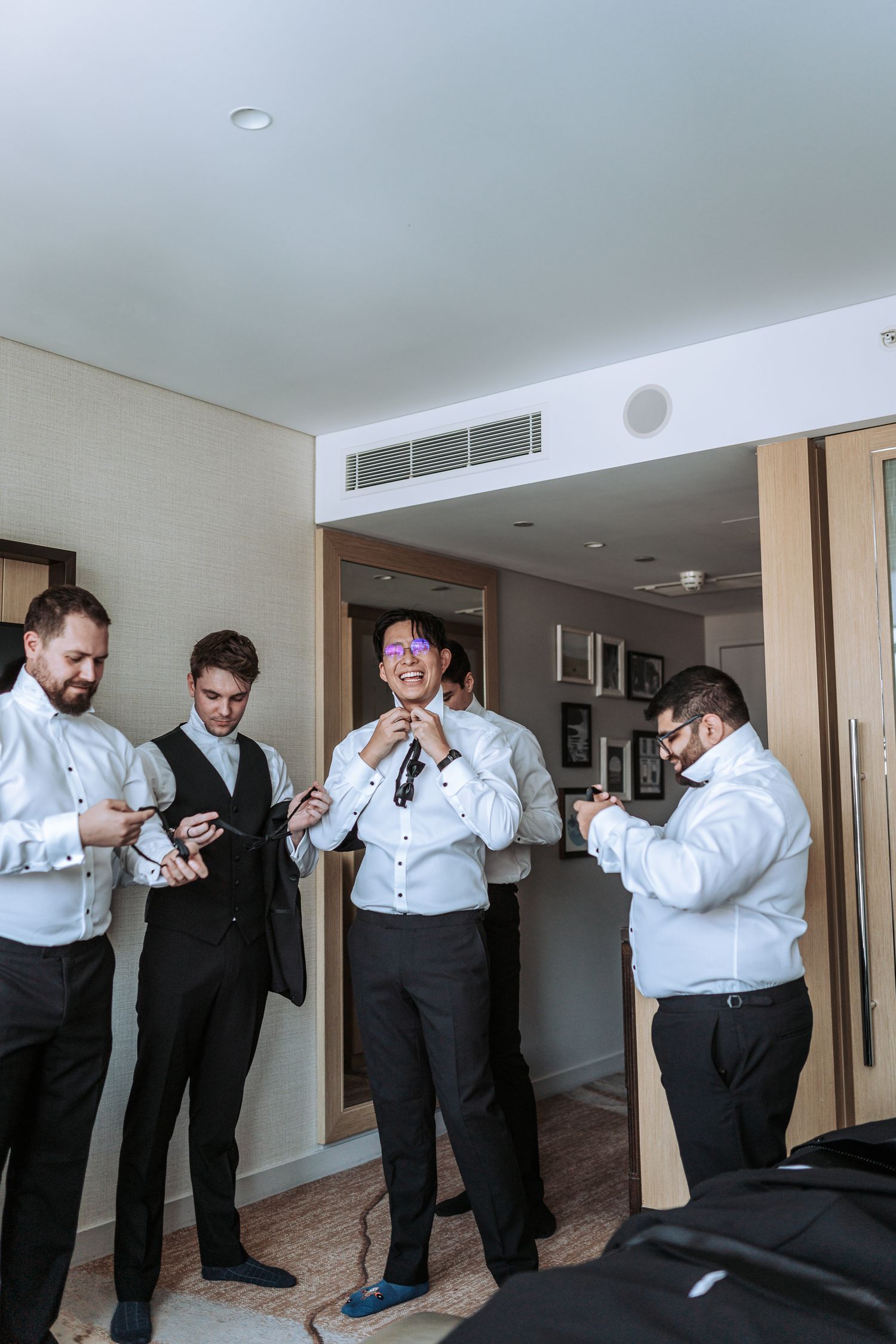 Group of groomsmen preparing and adjusting formal attire in hotel room.