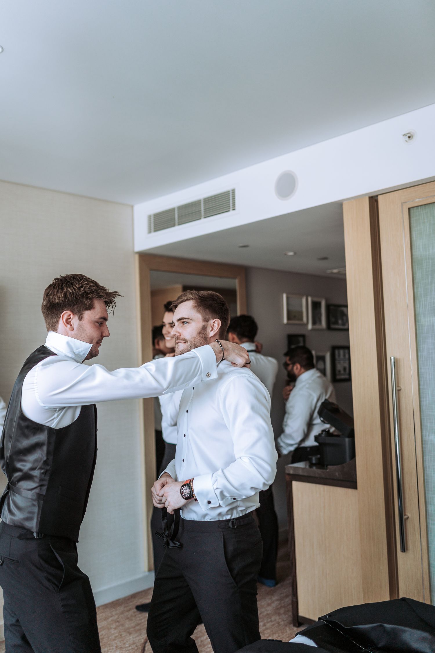 Wedding party members helping with formal attire adjustments in hotel room.
