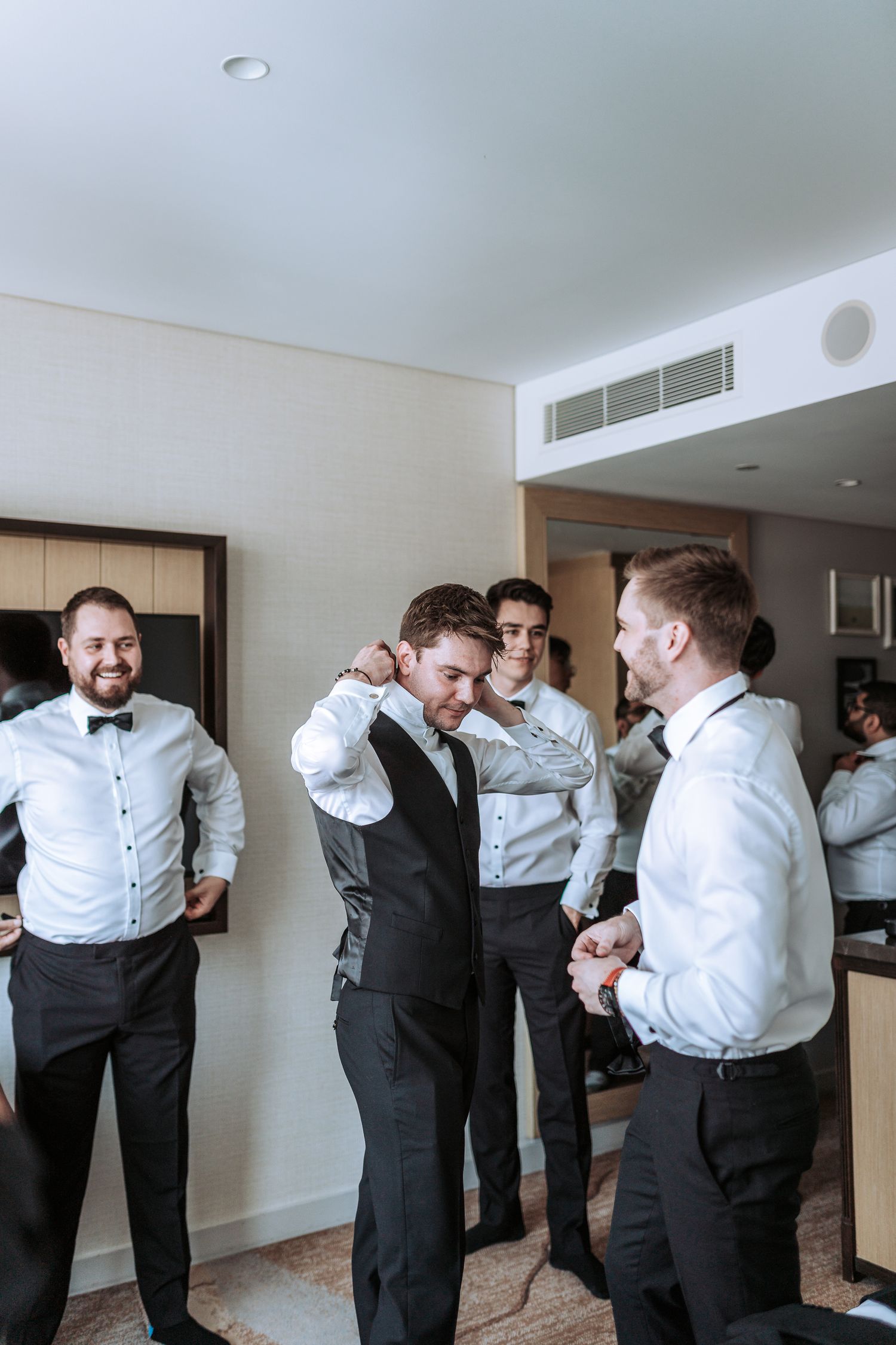Groomsmen in formal attire share a lighthearted moment while getting ready in a hotel room.