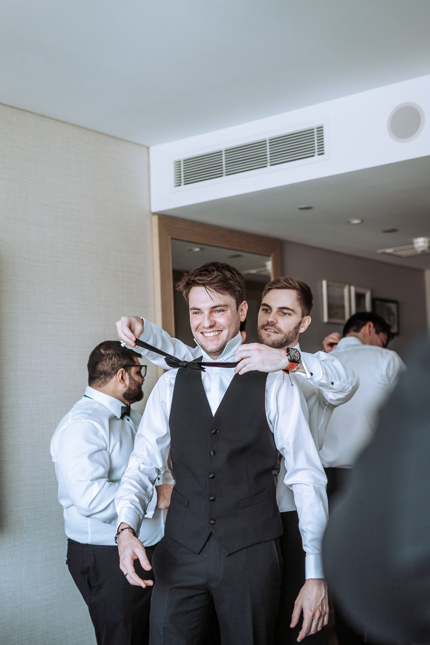 A group helps adjust a bowtie on a groomsman wearing a formal vest during wedding preparations.