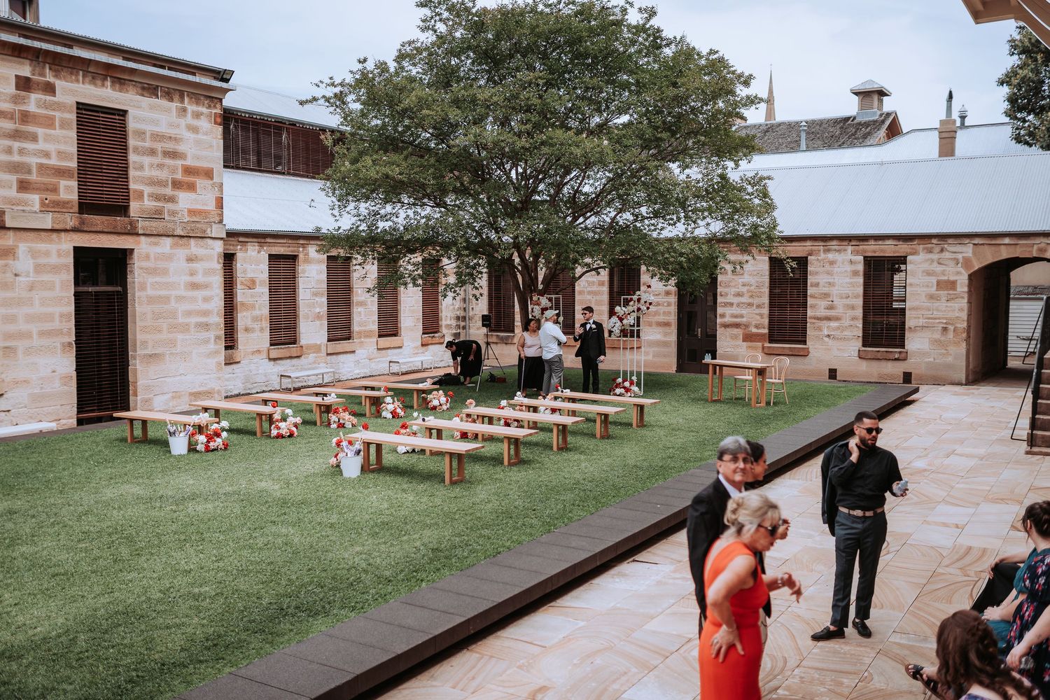 Outdoor courtyard wedding venue with wooden benches and stone architecture under large trees.