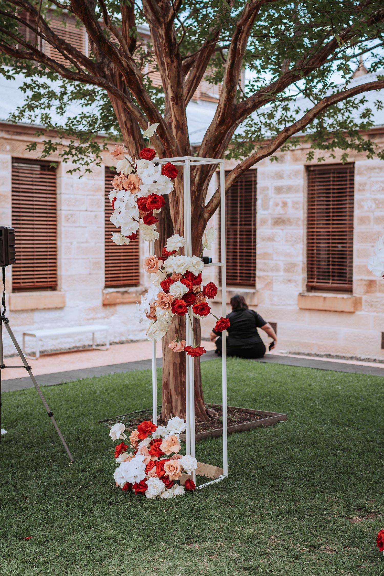 Modern white geometric arch decorated with red and white flowers against a large tree.