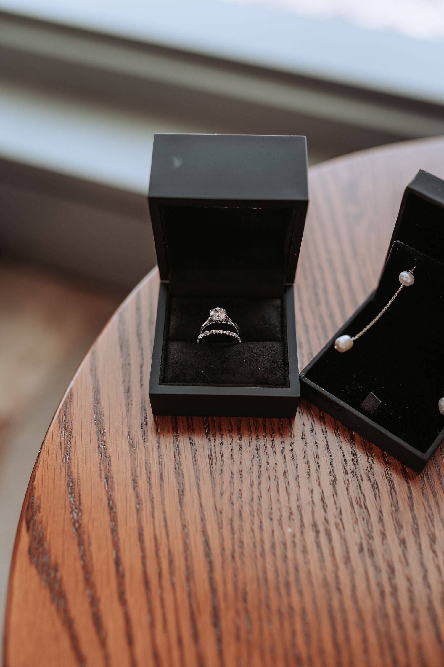 Diamond engagement ring and jewelry displayed in black velvet boxes on wooden surface.