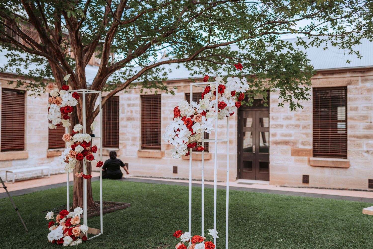 Outdoor wedding ceremony setup with white geometric arches adorned with red and white floral arrangements.