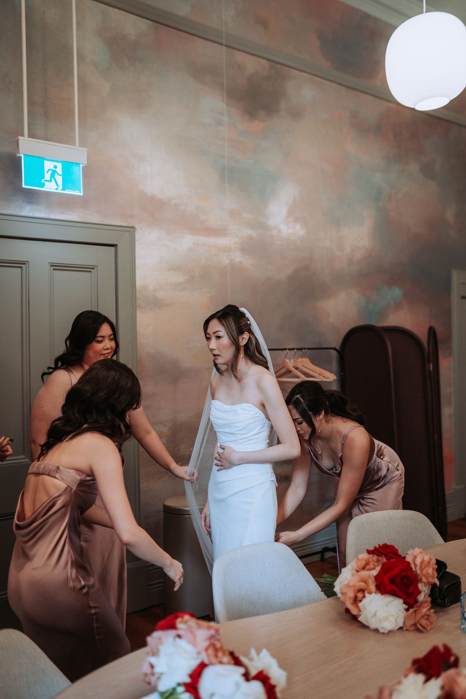 Bride in white dress getting help with final adjustments while bridesmaids in blush dresses assist.