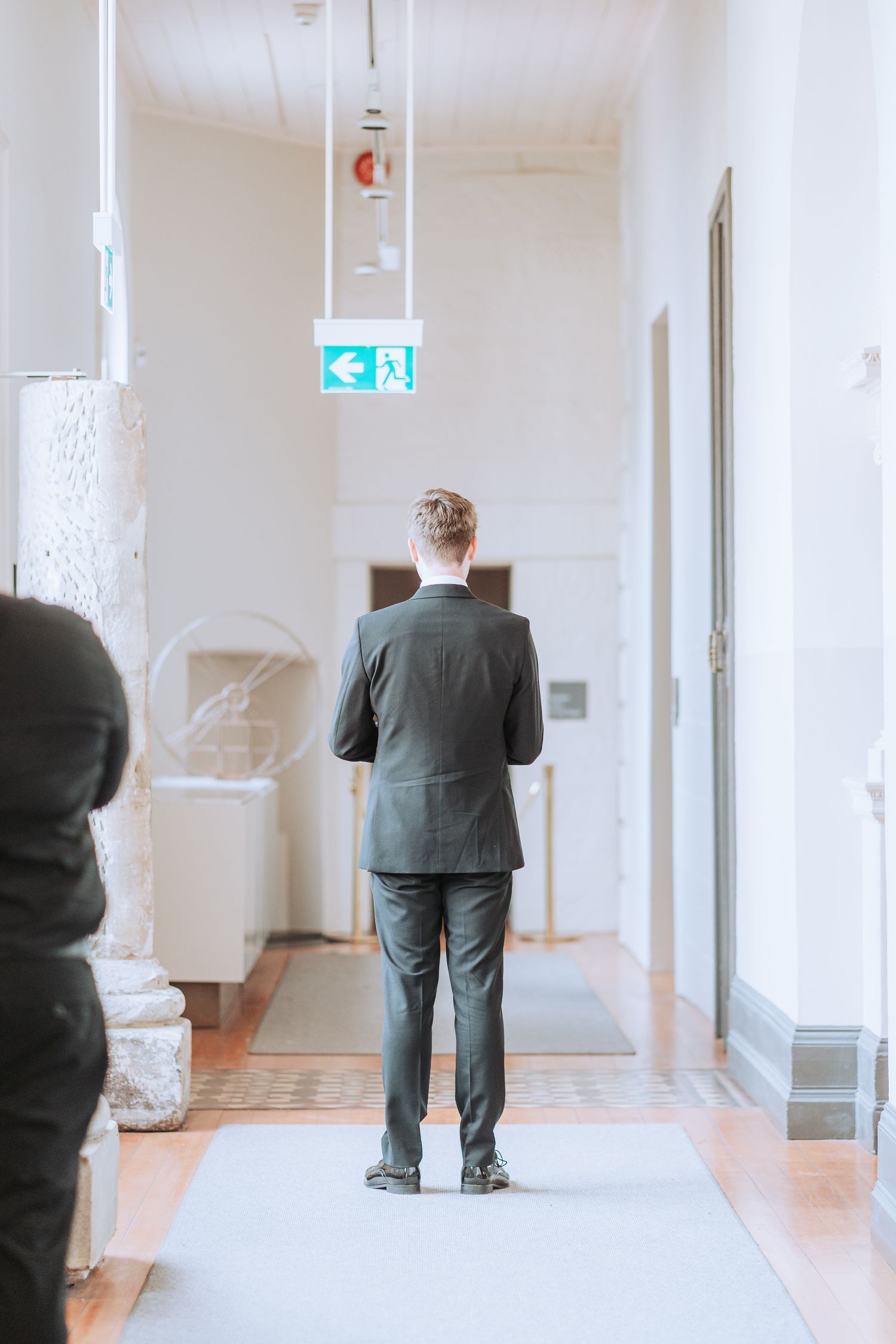 Person in formal suit standing alone in white hallway with emergency exit sign ahead.