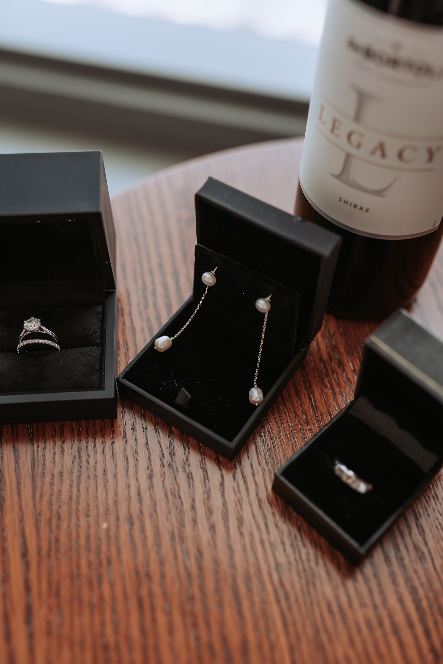 Elegant jewelry boxes containing rings and earrings displayed next to a Legacy wine bottle on a wooden surface.