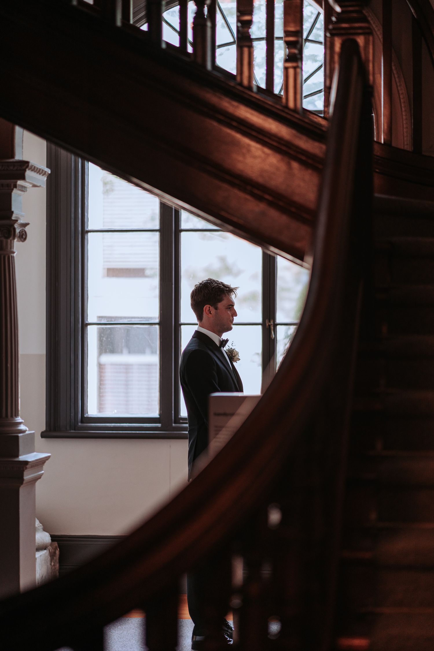 Groom standing by window with curved wooden staircase in foreground.