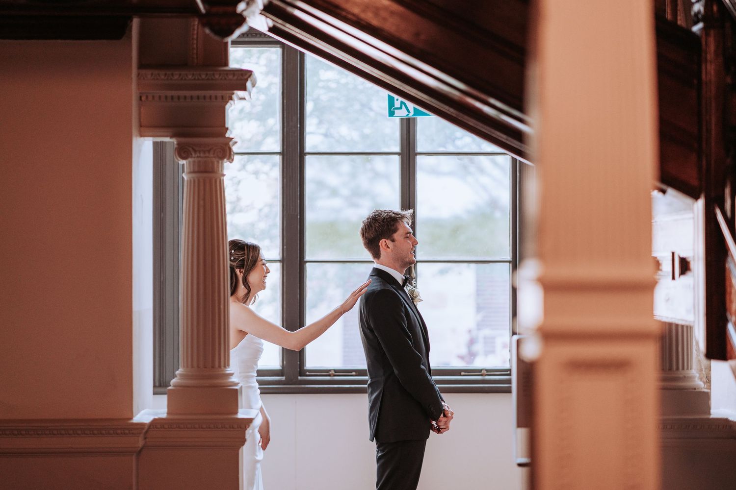 A formal couple stands back-to-back in an elegant venue with white columns.