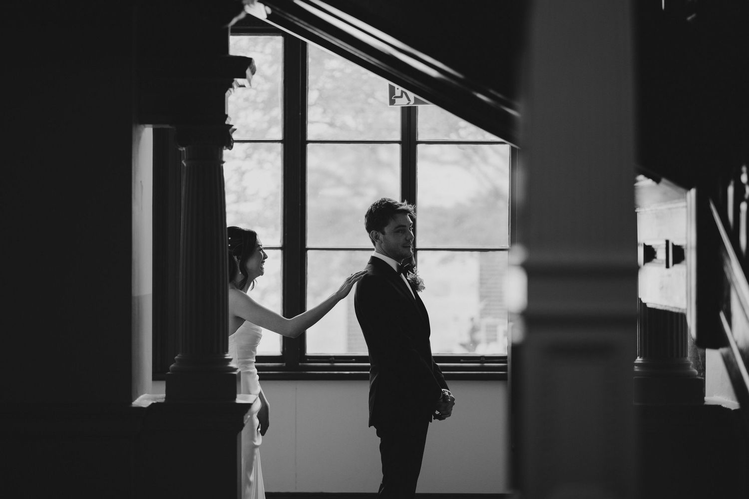 Two silhouettes stand in dramatic lighting by a large window creating a moody black and white composition.