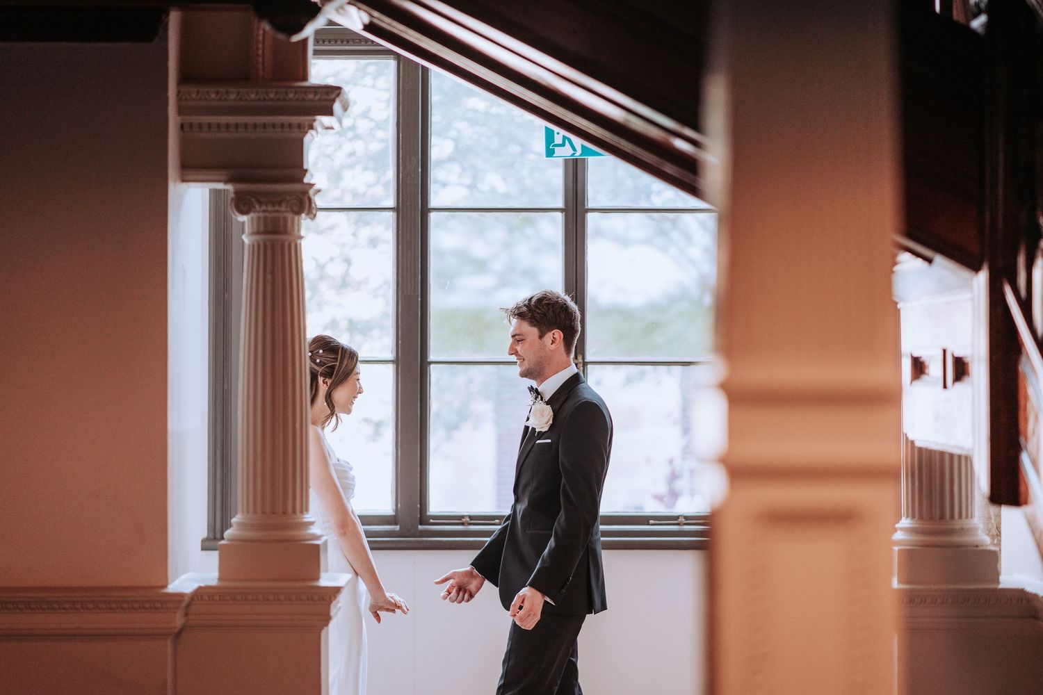 A couple stands facing each other in an elegant room with ornate columns and large windows.
