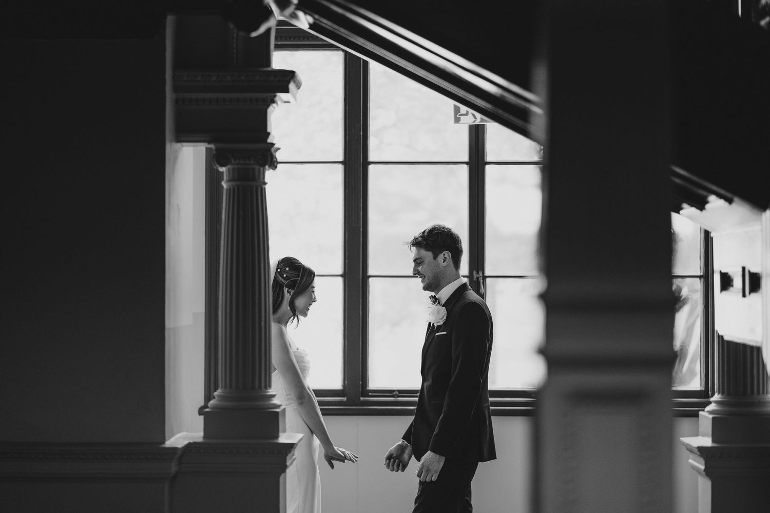 A black and white photo captures a couple standing between architectural columns near a window.