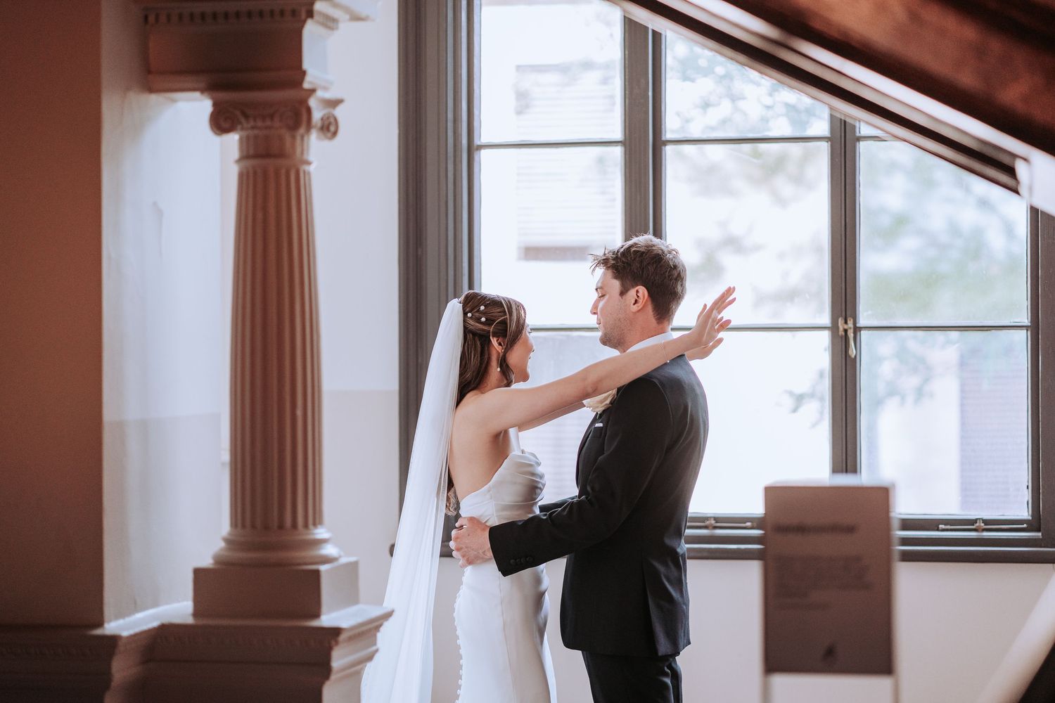 A couple embraces in an intimate moment by large windows and ornate columns.