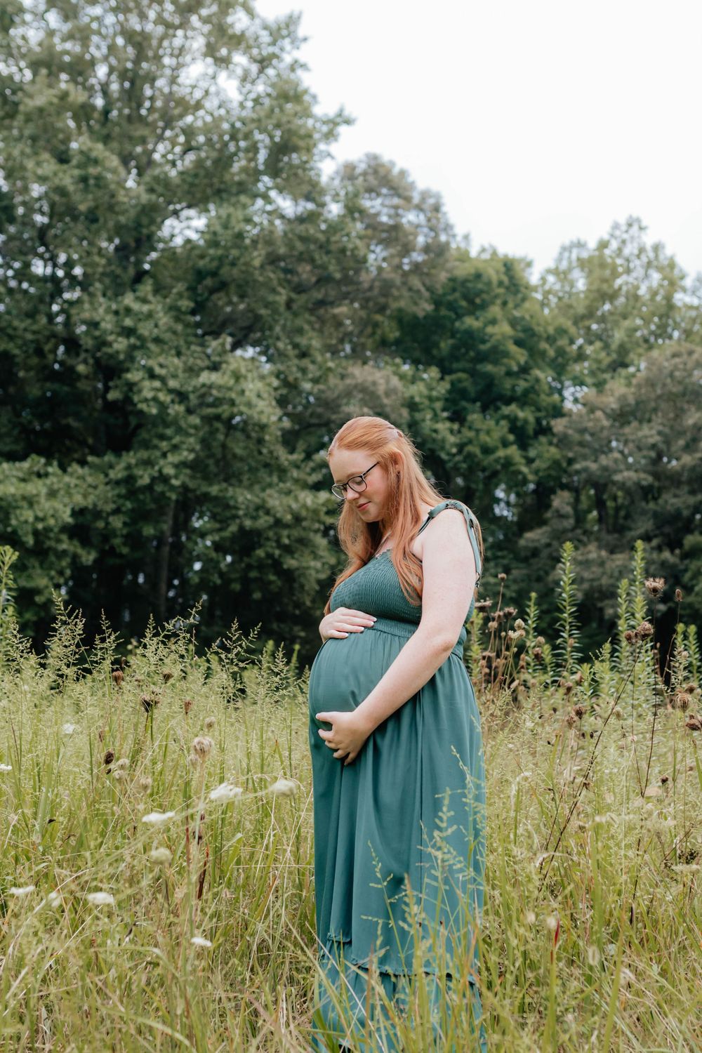 An expecting mother embracing in a serene meadow with tall grass and wildflowers.
