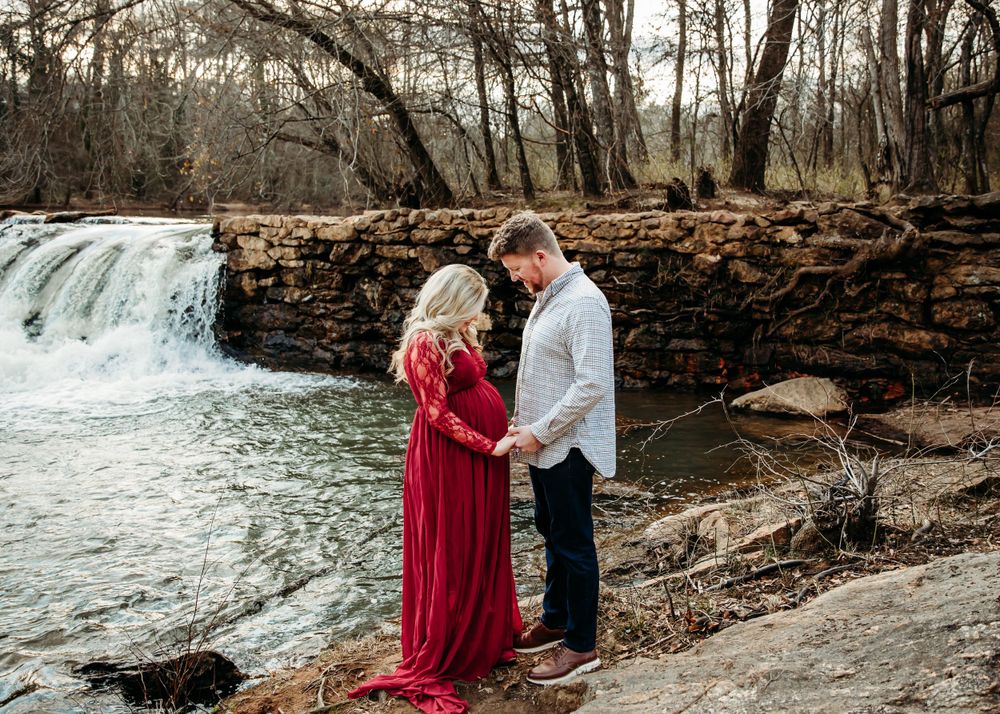 A couple poses near a scenic waterfall and stone wall during a romantic outdoor photoshoot.