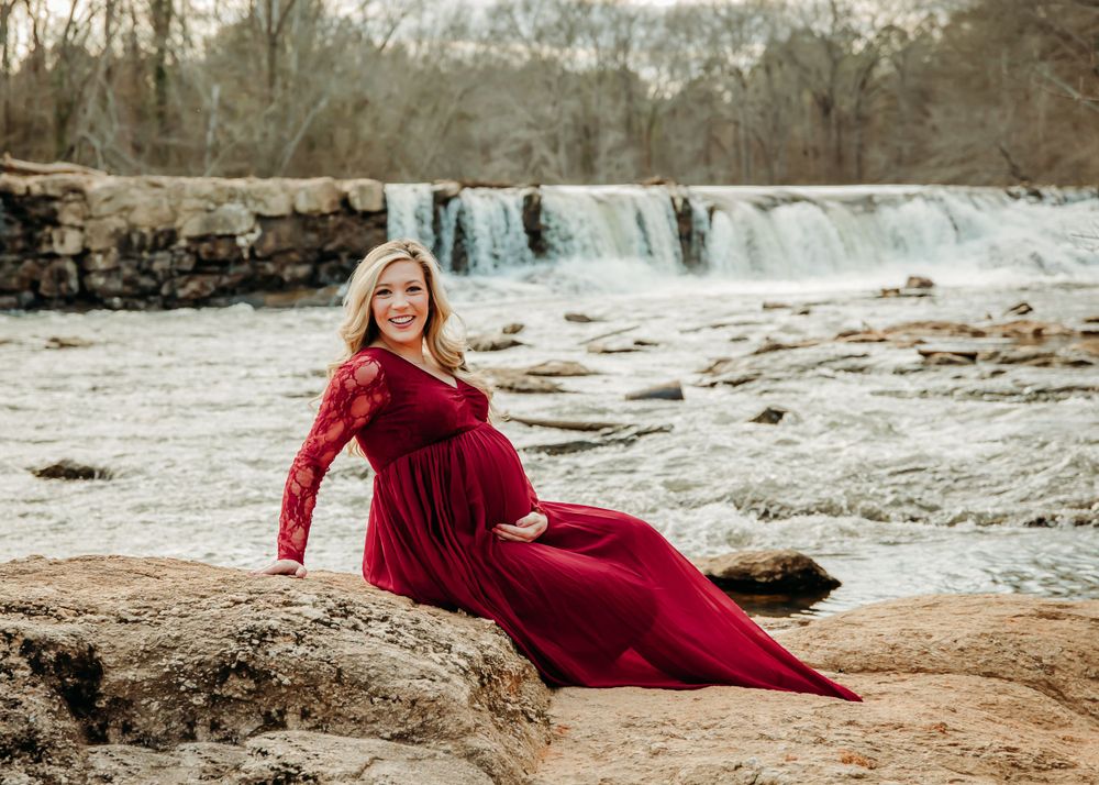 Woman in red dress poses near rushing waterfall on rocky shore.
