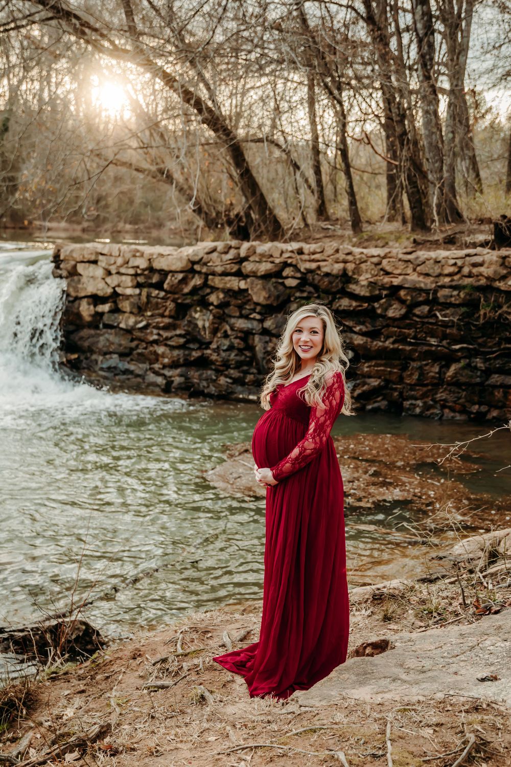 A burgundy dress flows gracefully by a stone wall and waterfall during a fall photoshoot in nature.