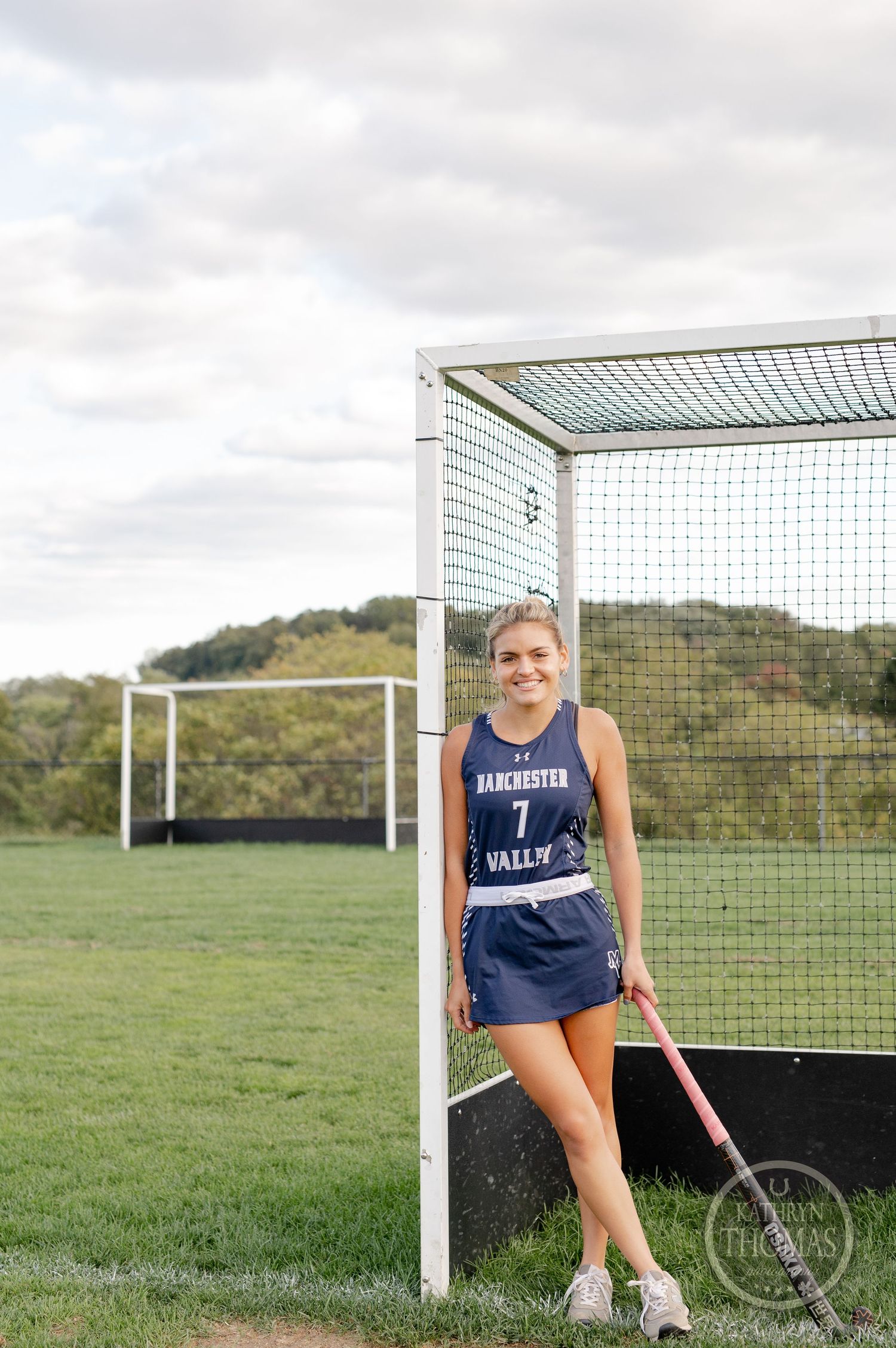 Field hockey athlete stands by goal post wearing navy jersey and holding stick.