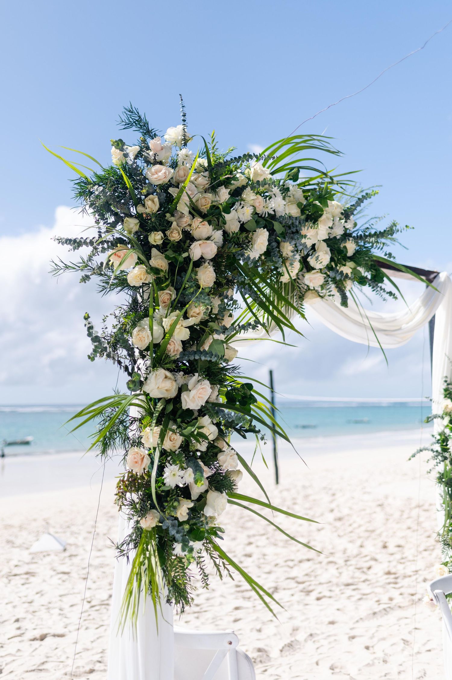White roses and tropical greenery adorn a wedding arch on a sandy beach with blue ocean and sky backdrop.