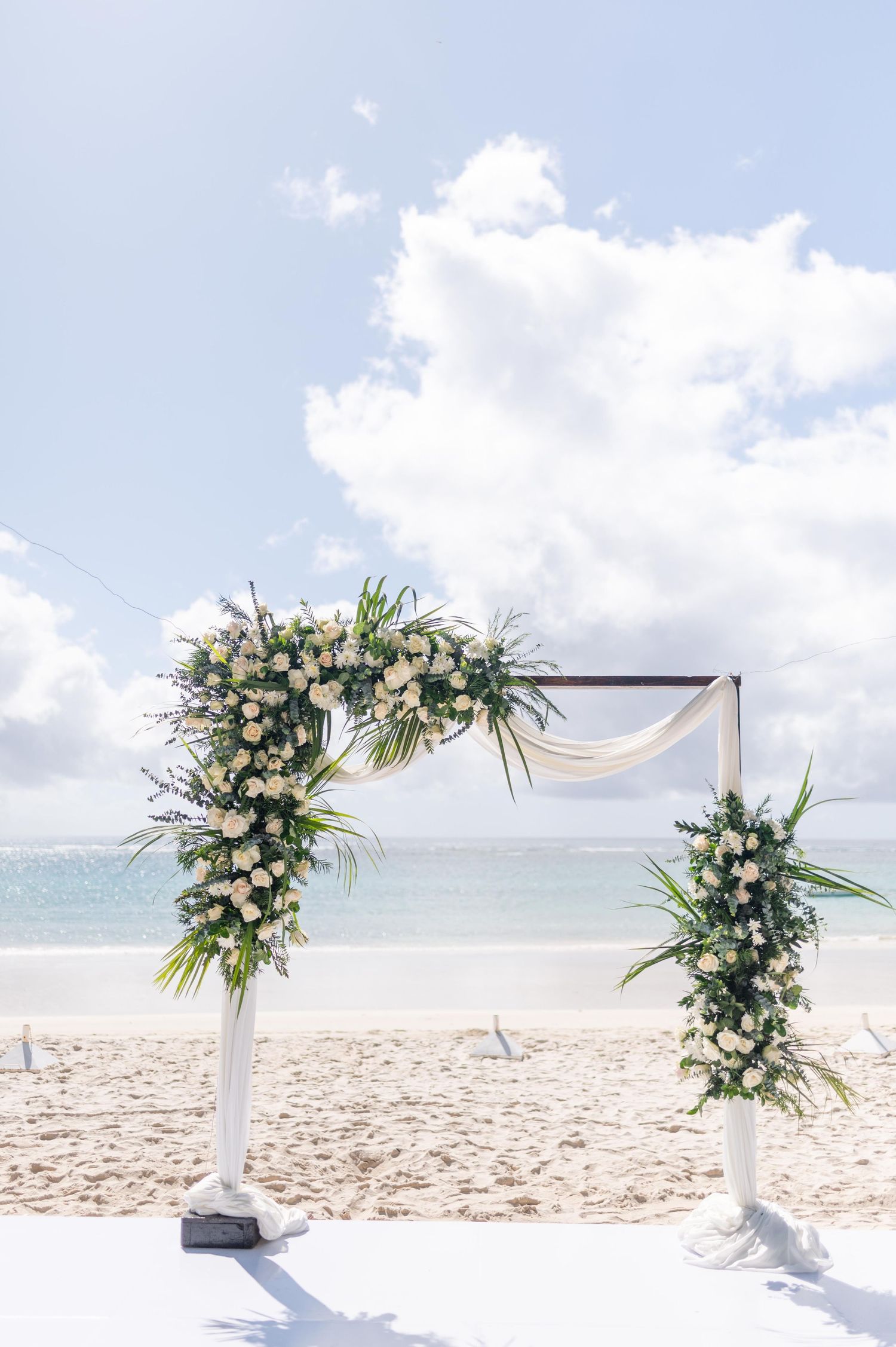 Elegant beach wedding arch adorned with white roses and tropical greenery, set against turquoise ocean and blue sky.