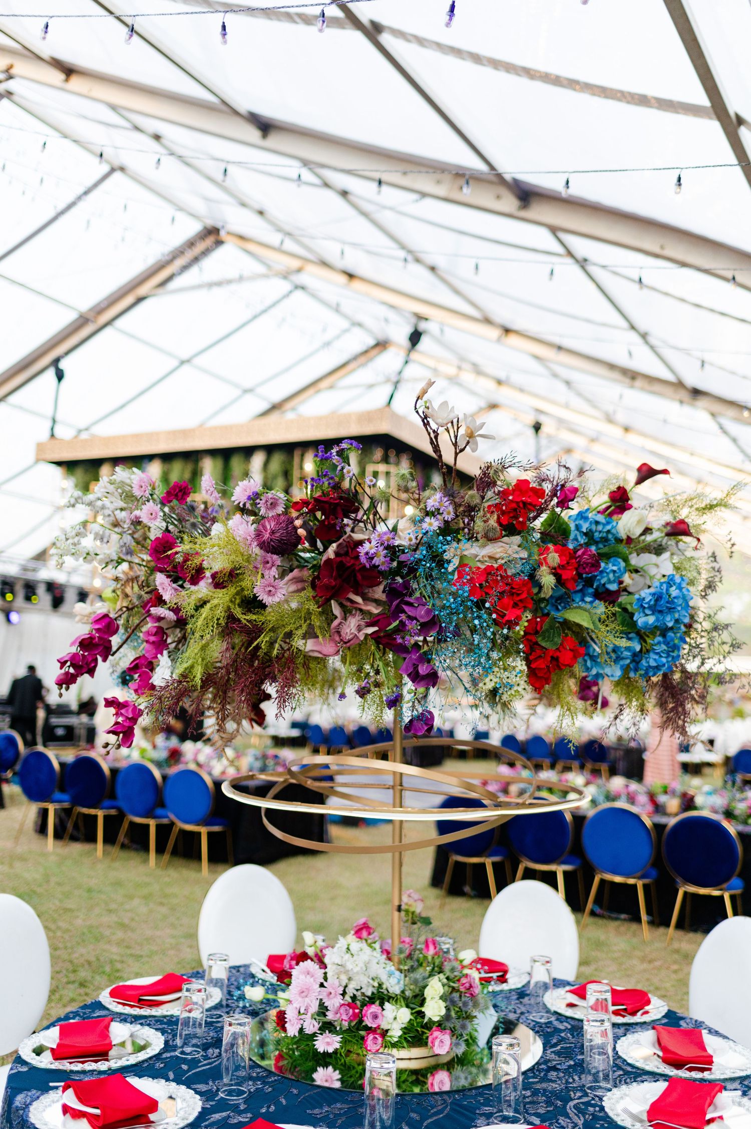 Elegant wedding reception under a clear tent with vibrant floral centerpieces in red, blue, and purple atop gold stands.