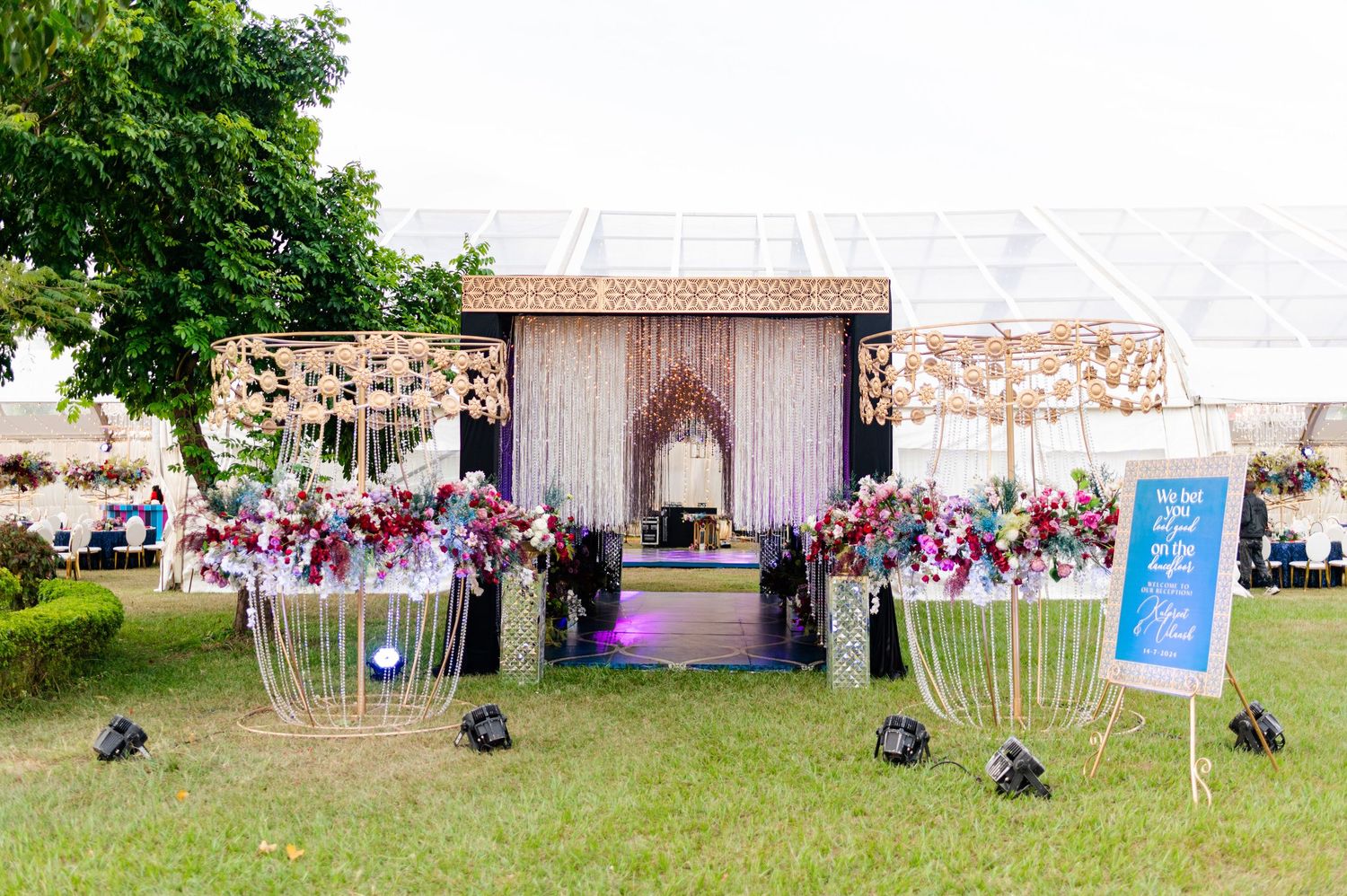 Elegant outdoor wedding backdrop with floral arch, draped curtains, and geometric gold stands on green lawn.