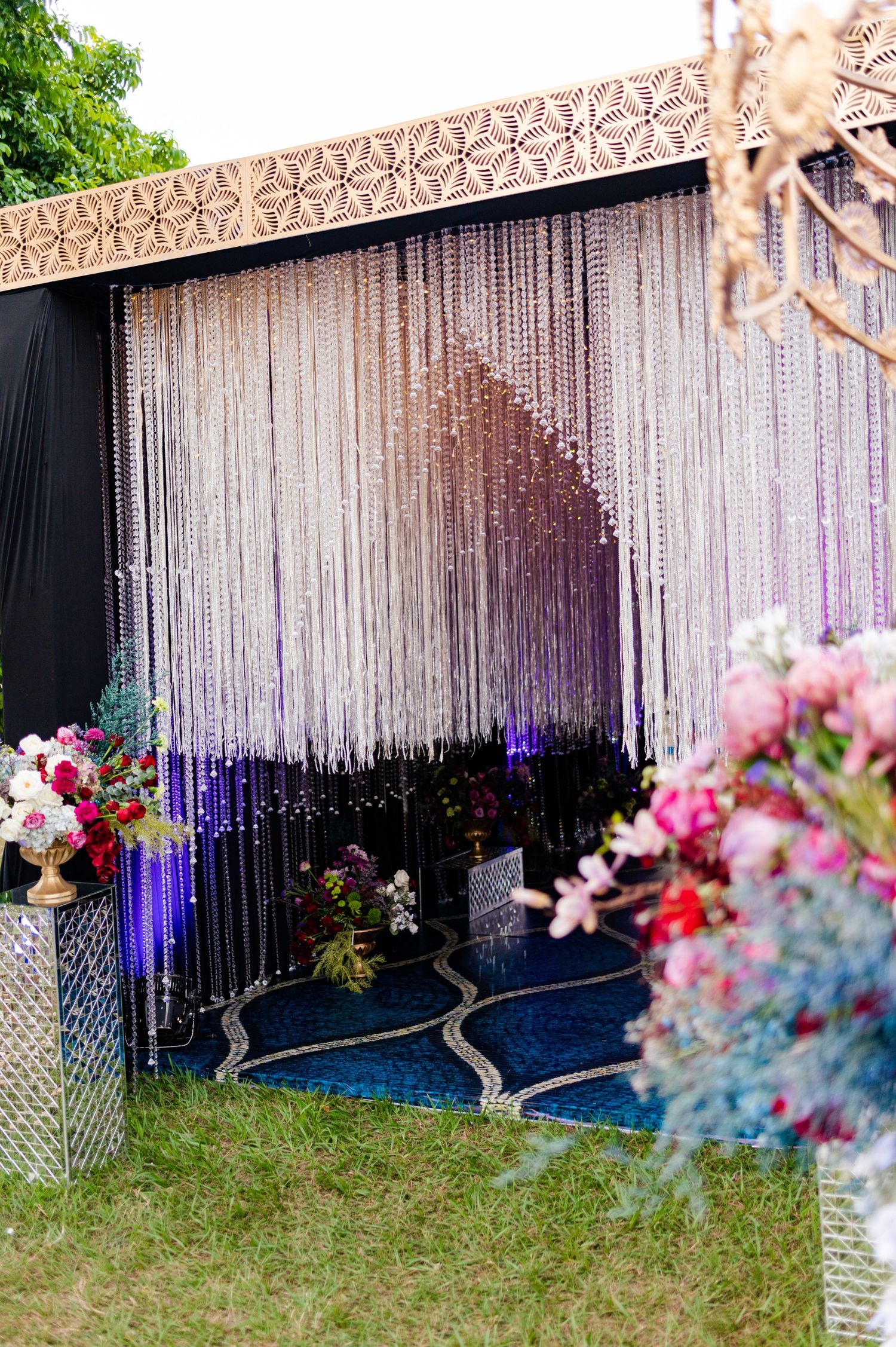 Glamorous wedding backdrop featuring cascading crystal bead curtains, ornate gold trim, and colorful floral arrangements.